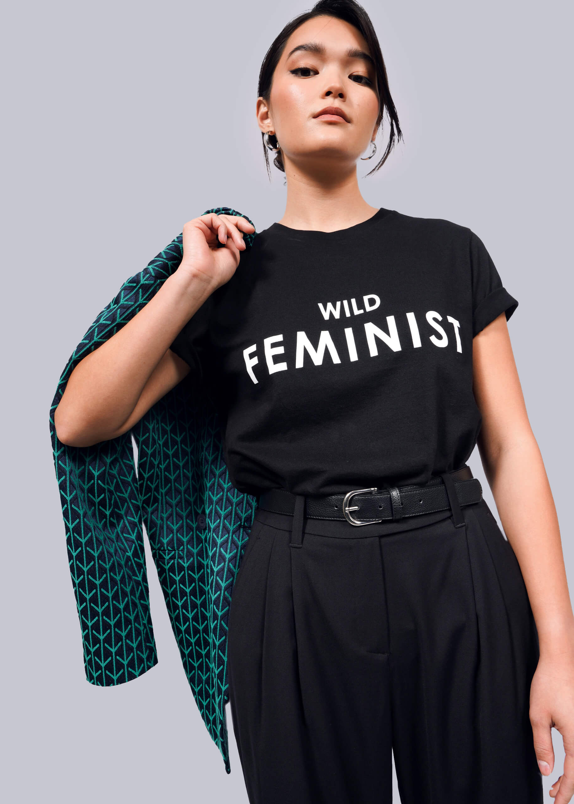 A woman confidently poses in The Wild Feminist® Tee, a black unisex fit T-shirt with WILD FEMINIST in bold white letters, made from pre-shrunk 100% cotton for lasting comfort. She holds a jacket over her shoulder and pairs it with high-waisted black pants against a plain white background.