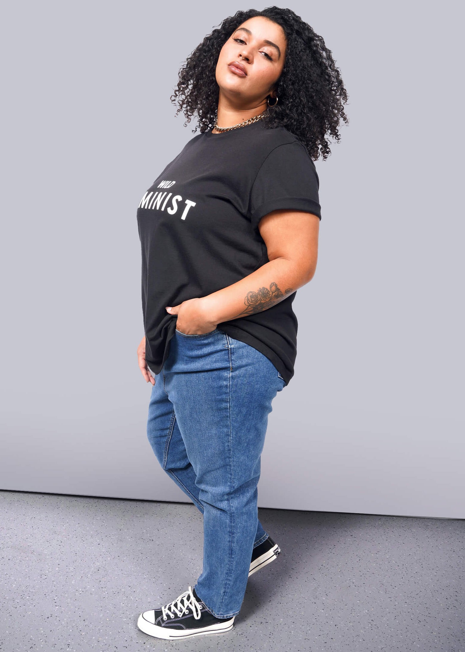 1X=A person with curly hair confidently wears The Wild Feminist® Tee, a black cotton unisex shirt with Feminist printed on it. Paired with blue jeans and black sneakers, they pose boldly against a plain white background.