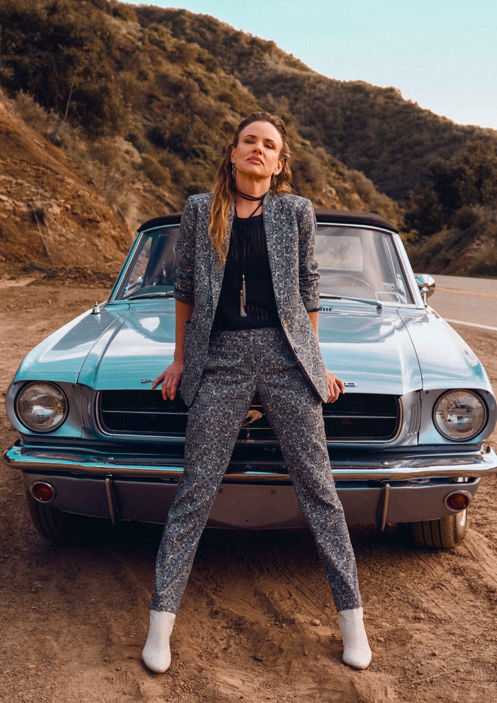 Juliette Lewis, wearing her teal bandana print suit, stands in front of vintage car in desert environment.