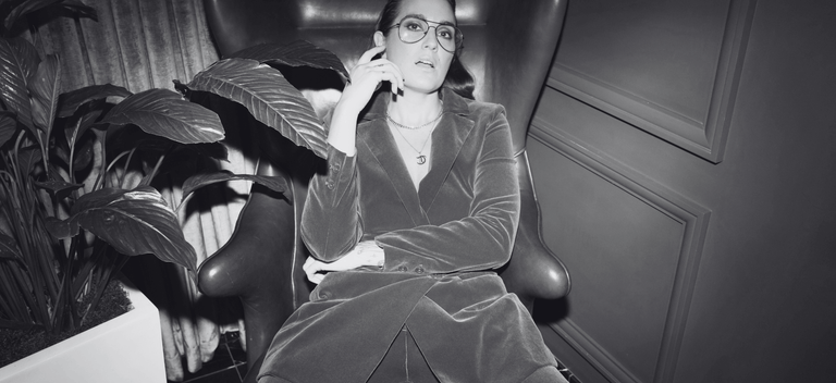 Black and white photo. Model is sitting in leather chair wearing velvet blazer in office setting.