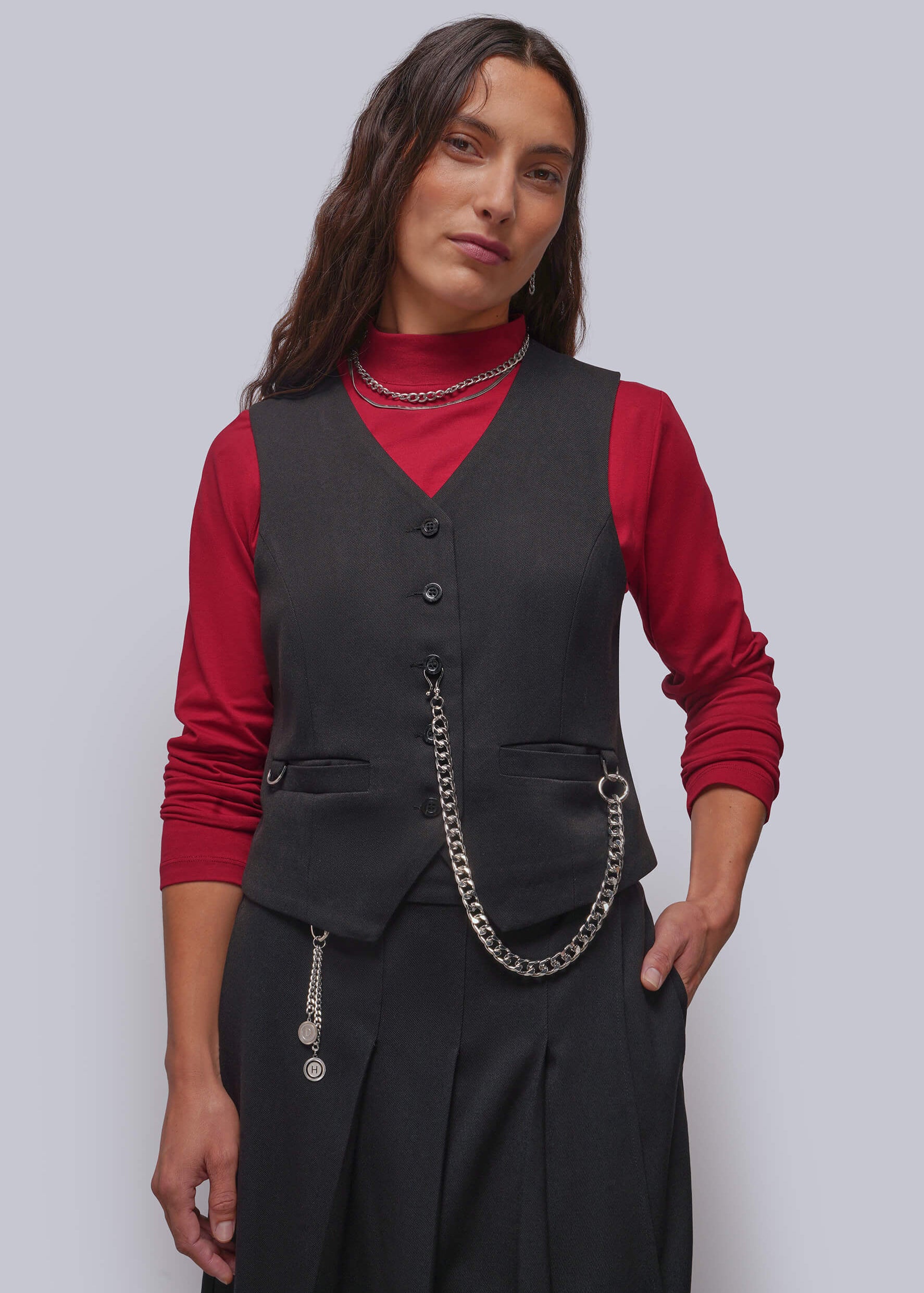 A woman modeling a black vest over a red long-sleeve top and black pleated skirt, accessorized with a silver chain belt and layered necklaces, against a solid gray studio background.