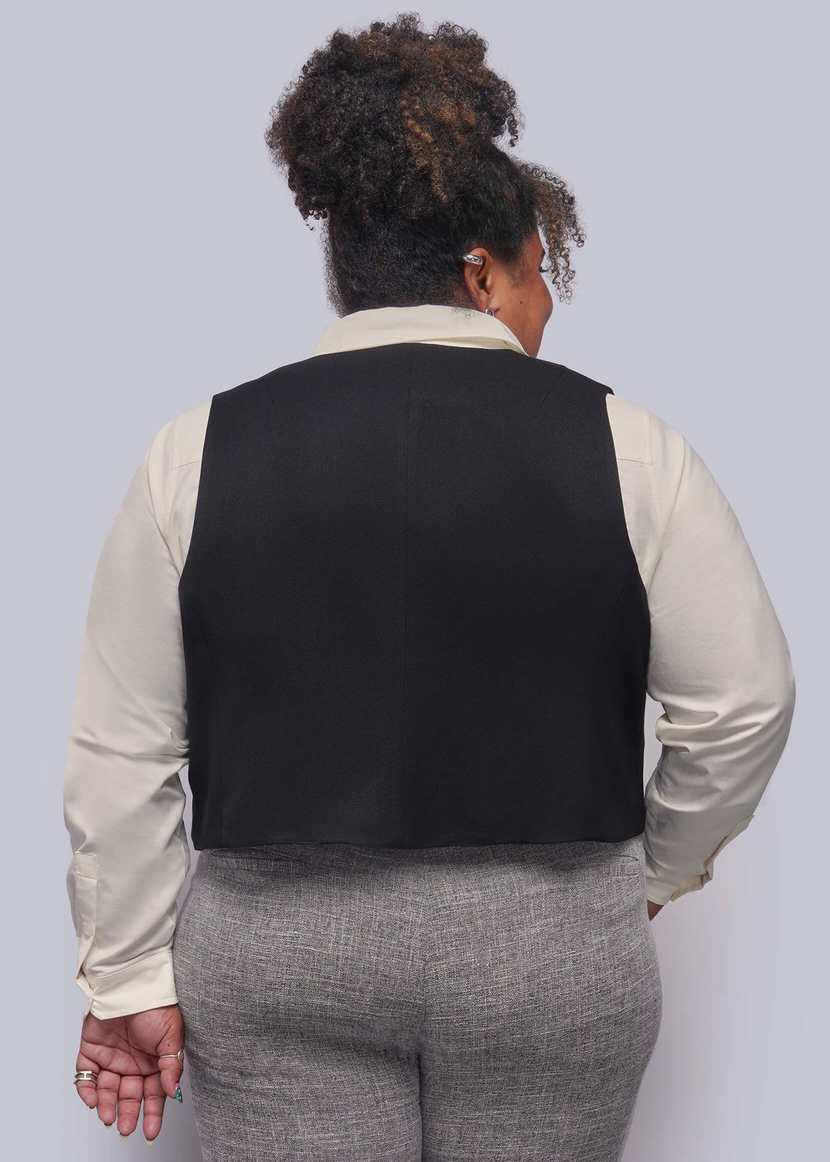 1X=A person shown from the back wearing a black vest over a cream long-sleeve shirt and gray textured pants, against a plain light gray studio background.