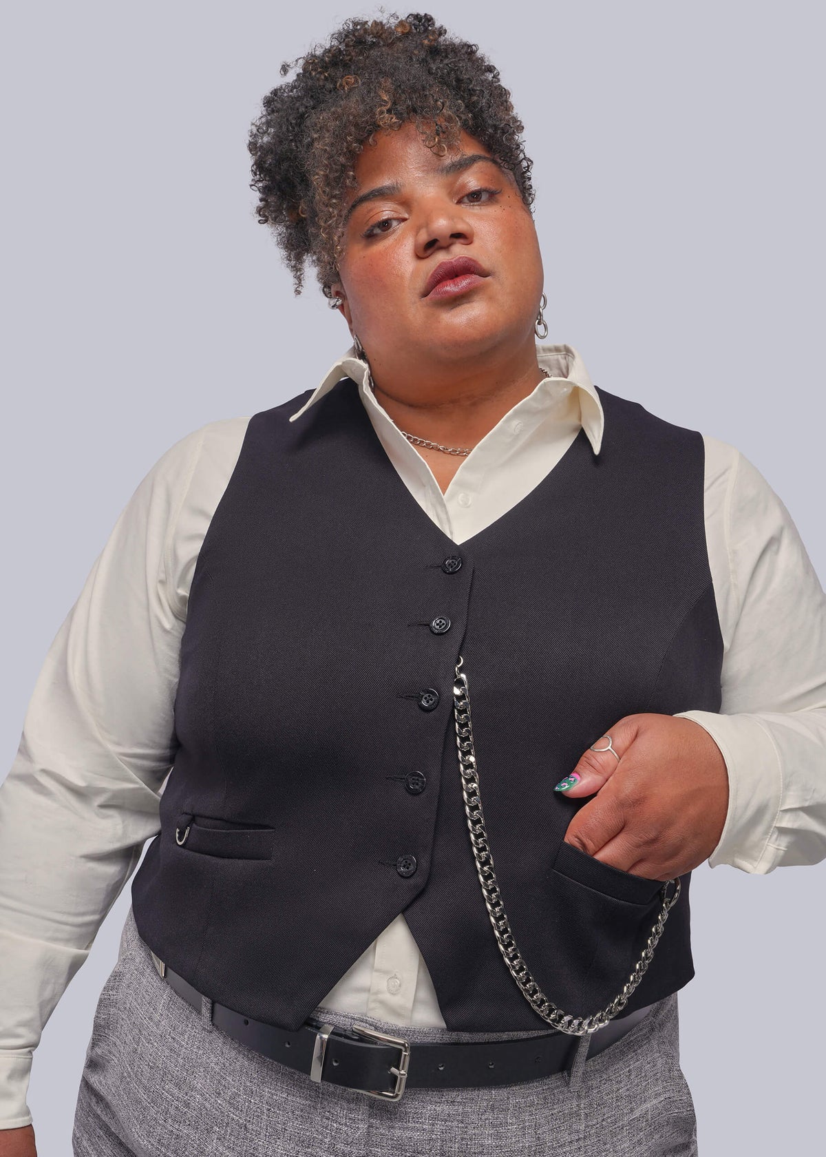1X=A plus-size model wearing a black button-front vest over a cream collared shirt, paired with gray trousers and a silver chain accessory, against a solid light gray background.