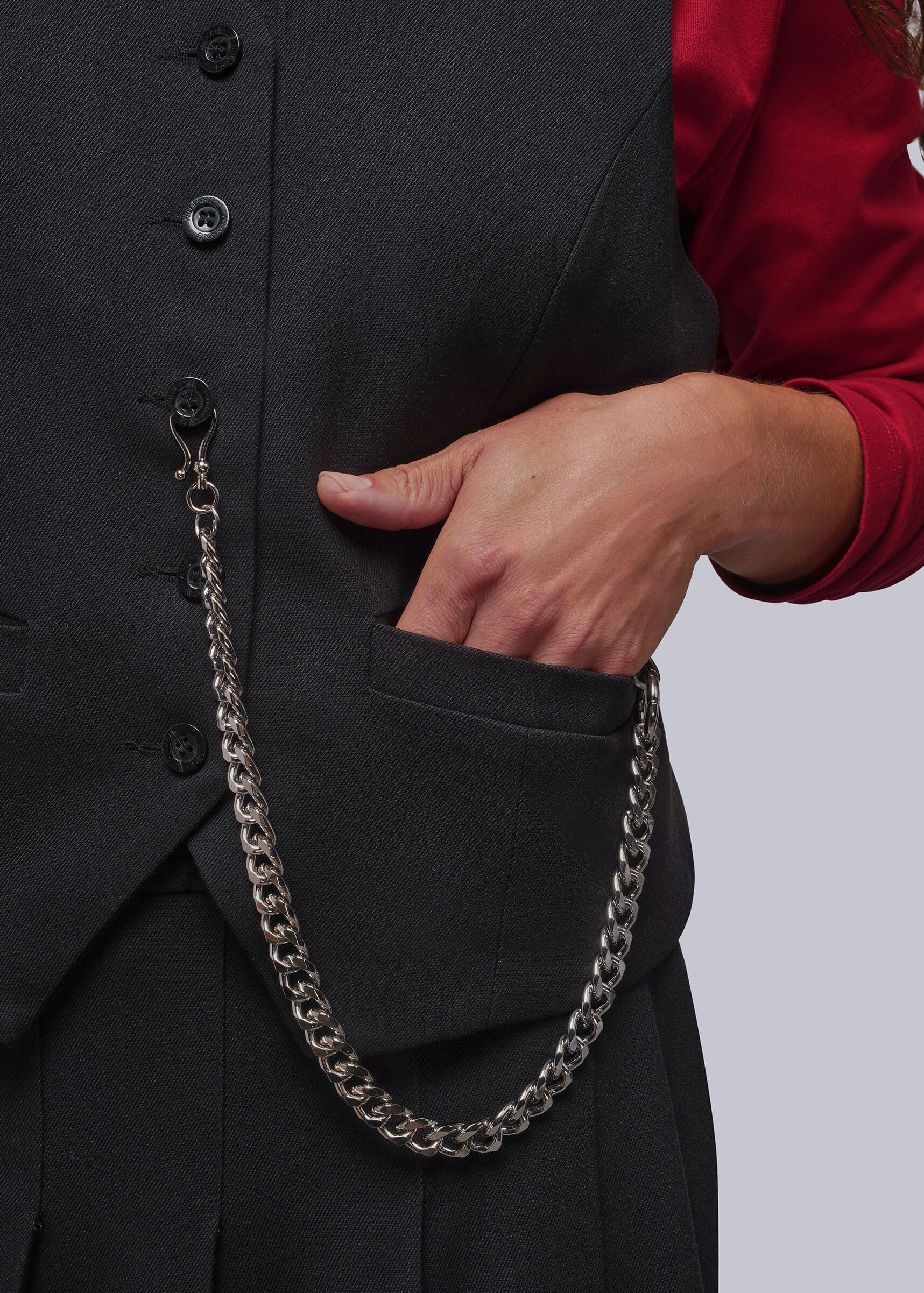A close-up of a person wearing a black vest and skirt, with a silver chain accessory draped from the vest pocket over a red long-sleeve top, against a light gray background.