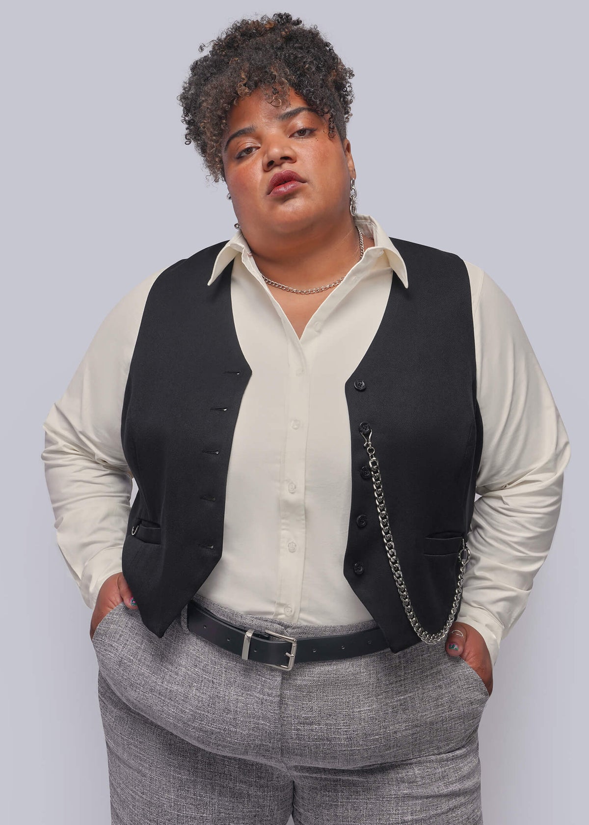 1X=A plus-size model wearing a black vest over a cream button-up shirt, paired with gray textured trousers and a black belt, standing against a solid light gray background.