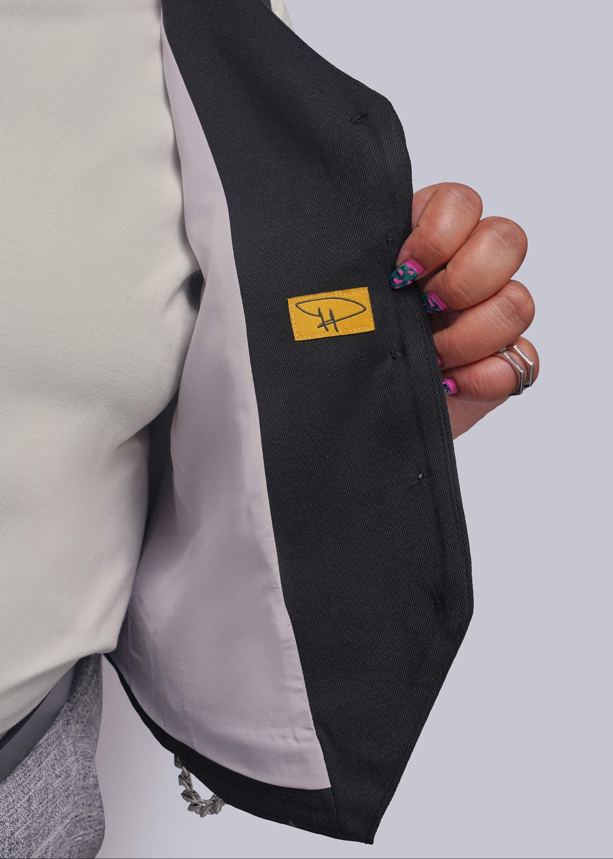 1X=Close-up of a person holding open a black vest to show the light gray lining and a small yellow brand patch with a stylized 'DH' logo.
