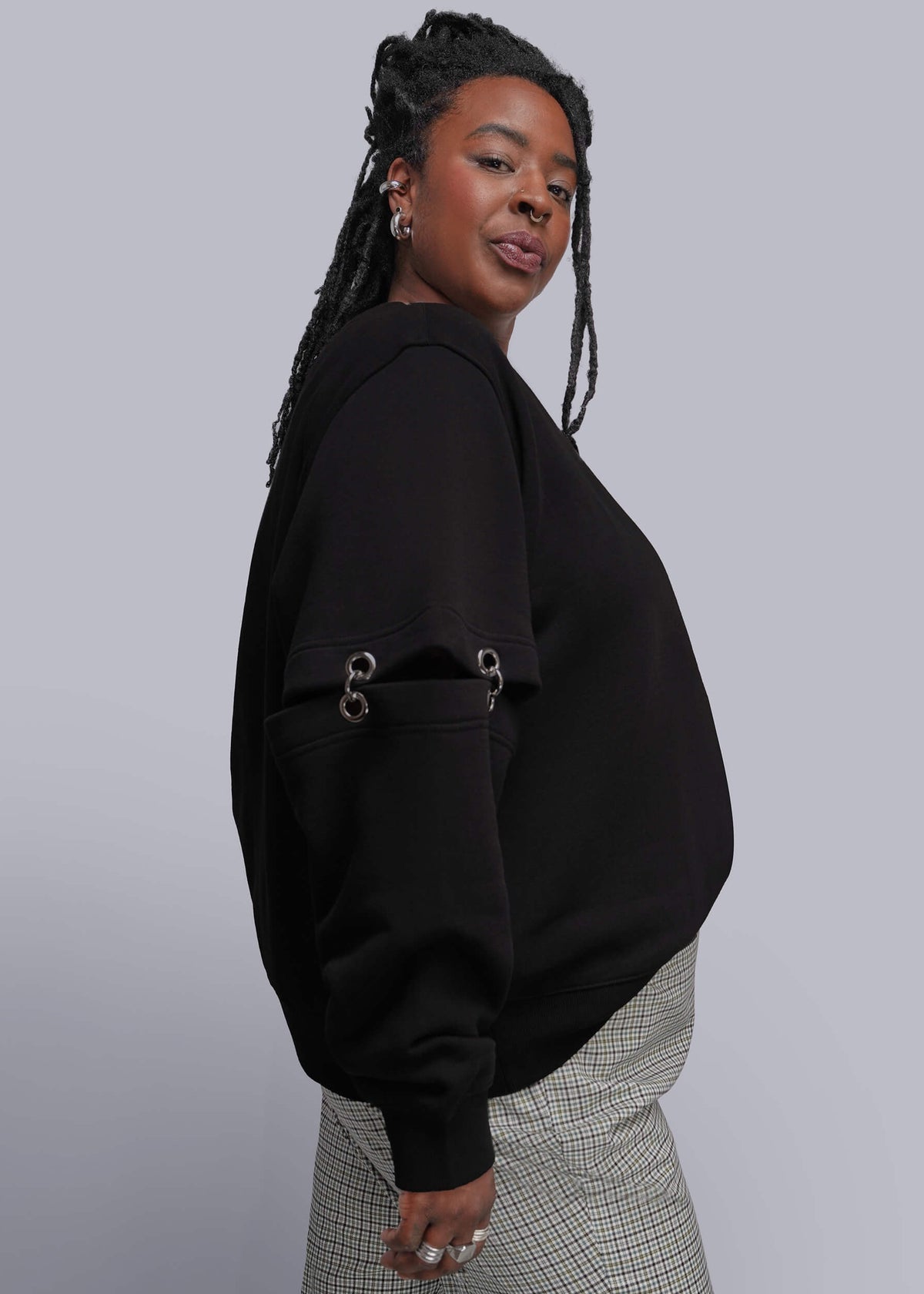 1X=A woman wearing a black cropped sweatshirt with silver grommet details on the sleeves, paired with gray and white checkered pants, photographed against a solid gray studio background.