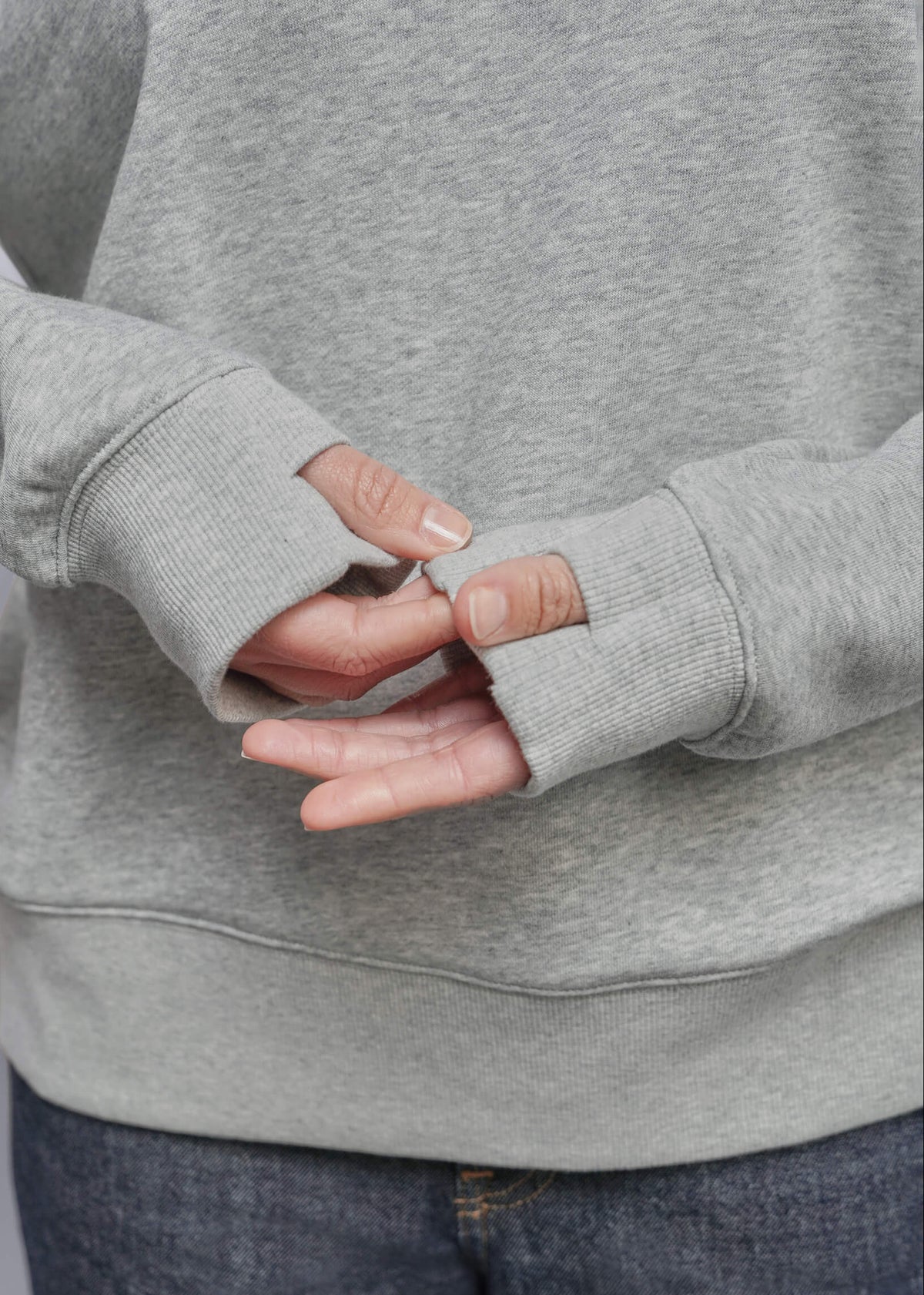 S=Close-up of a person wearing a light gray heather sweatshirt with thumbhole sleeves and dark blue jeans, showing the ribbed cuffs and hands.