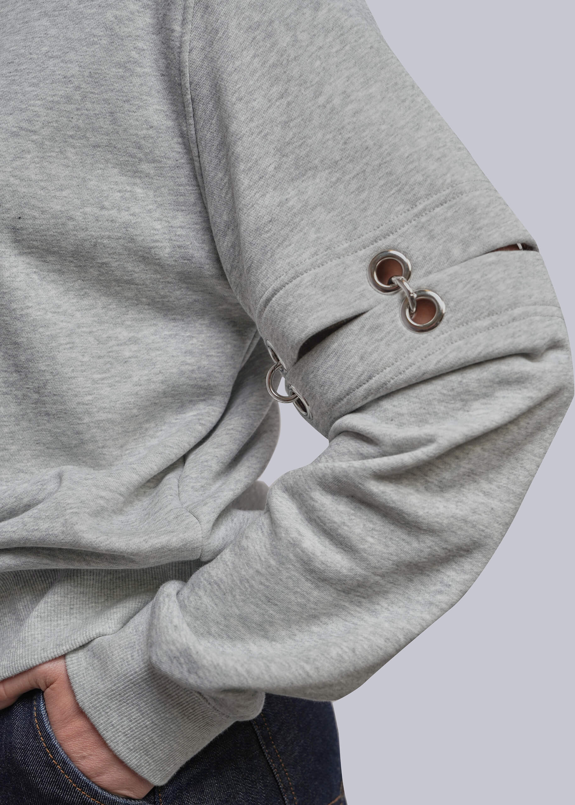 Close-up of a person wearing a heather gray long-sleeve sweatshirt with metal grommet details on the sleeve, paired with dark blue jeans against a light gray background.