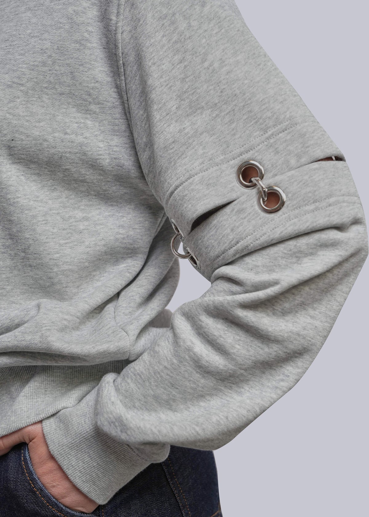 S=Close-up of a person wearing a heather gray long-sleeve sweatshirt with metal grommet details on the sleeve, paired with dark blue jeans against a light gray background.