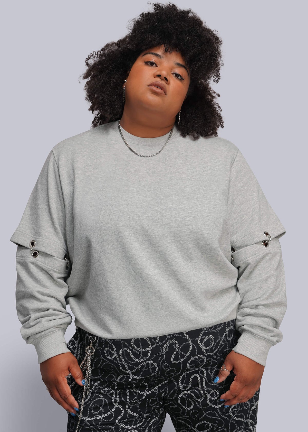 1X=A plus-size model with curly black hair wearing a light gray crewneck sweatshirt with grommet details on the sleeves and black pants with a white abstract pattern, set against a solid light gray background.