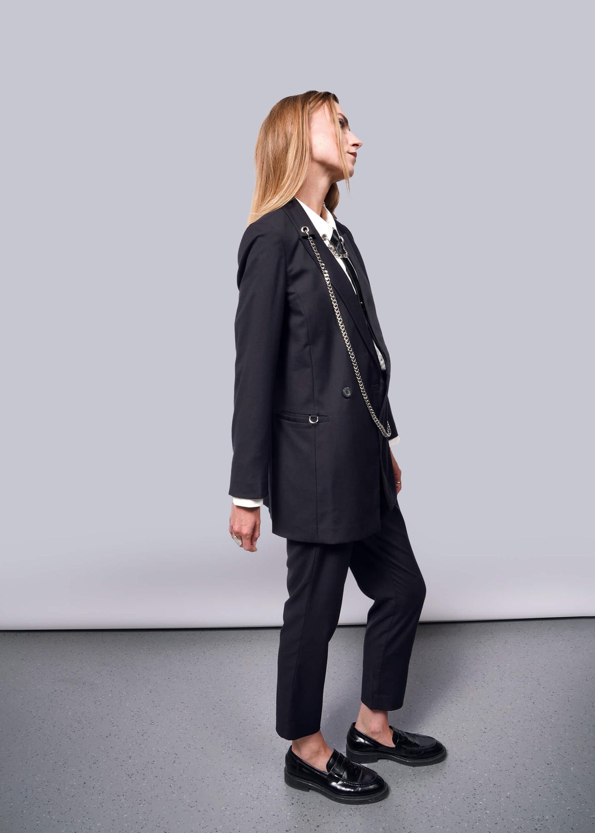 S=A person with long hair in profile against a plain background wears a Debbie Harry X Wildfang Double Breasted Blazer with metal detailing. They pair it with shiny black loafers, looking upward, their hands relaxed at their sides.