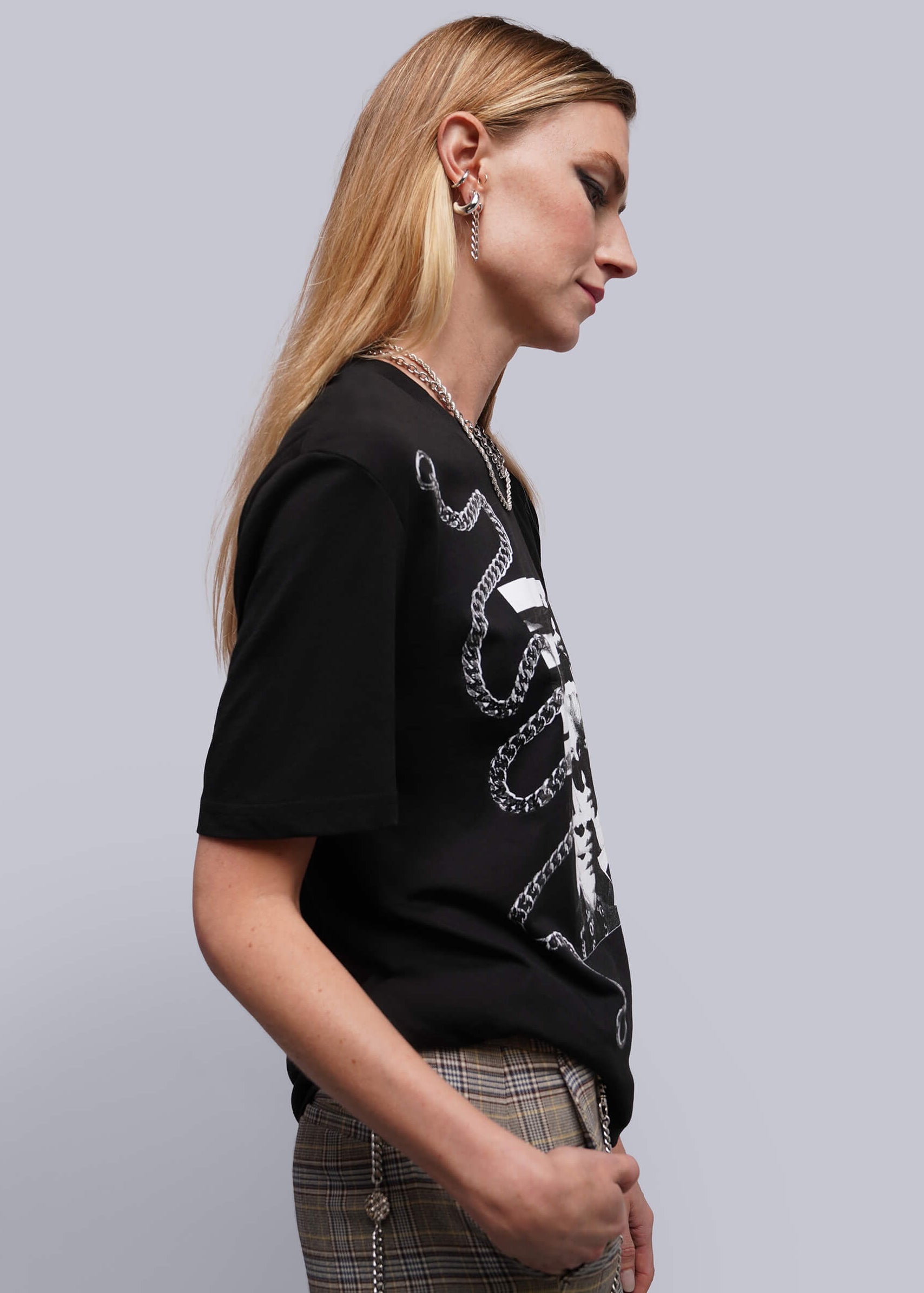 A woman in profile wearing a black short-sleeve graphic T-shirt with a white chain print, paired with plaid trousers, against a plain light gray studio background.