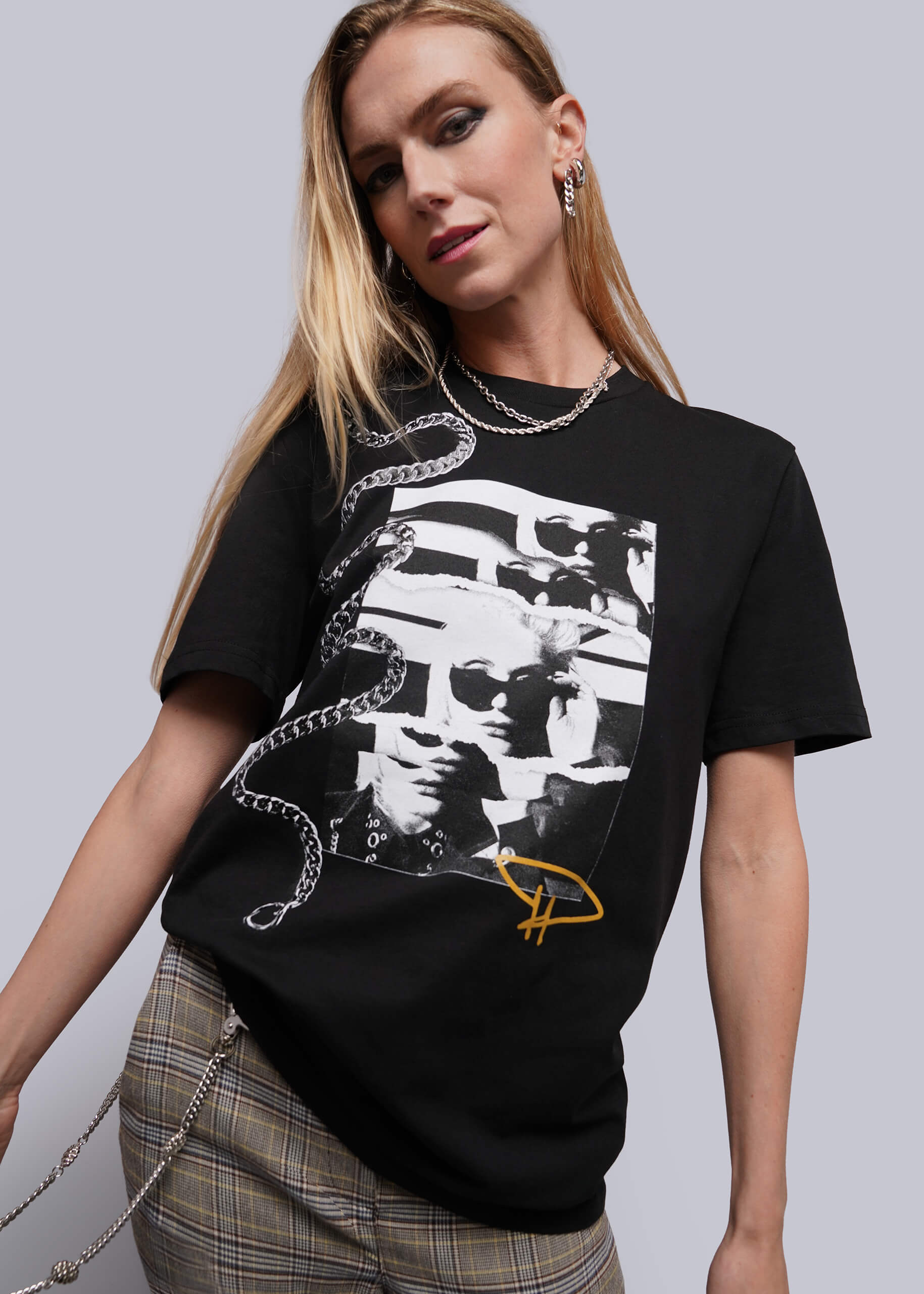A woman modeling a black graphic t-shirt featuring a white and black collage print of a face with chain graphics and a yellow logo, paired with plaid trousers against a solid gray background.