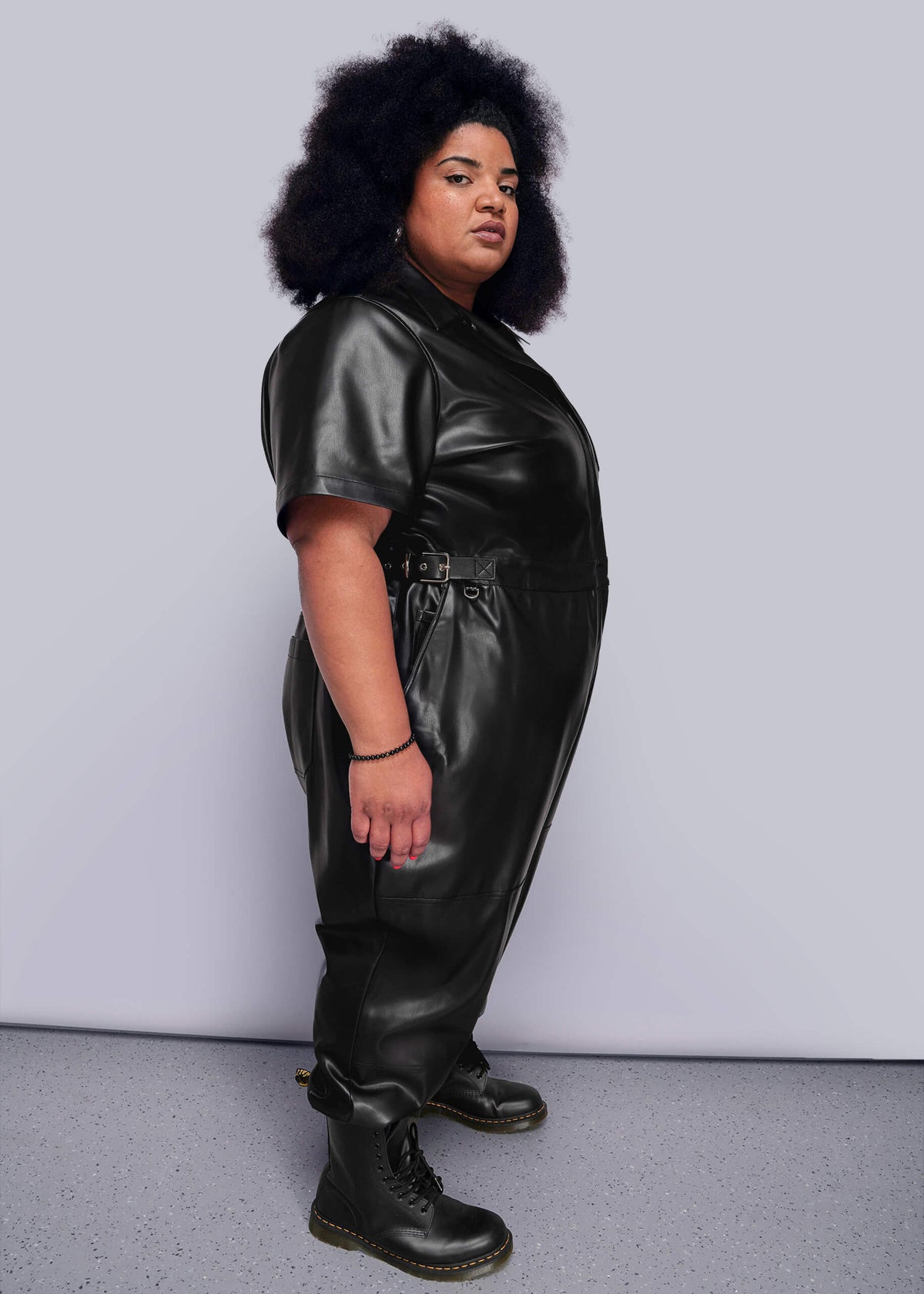 1X=Side view of person wearing a black vegan leather jumpsuit against a plain background