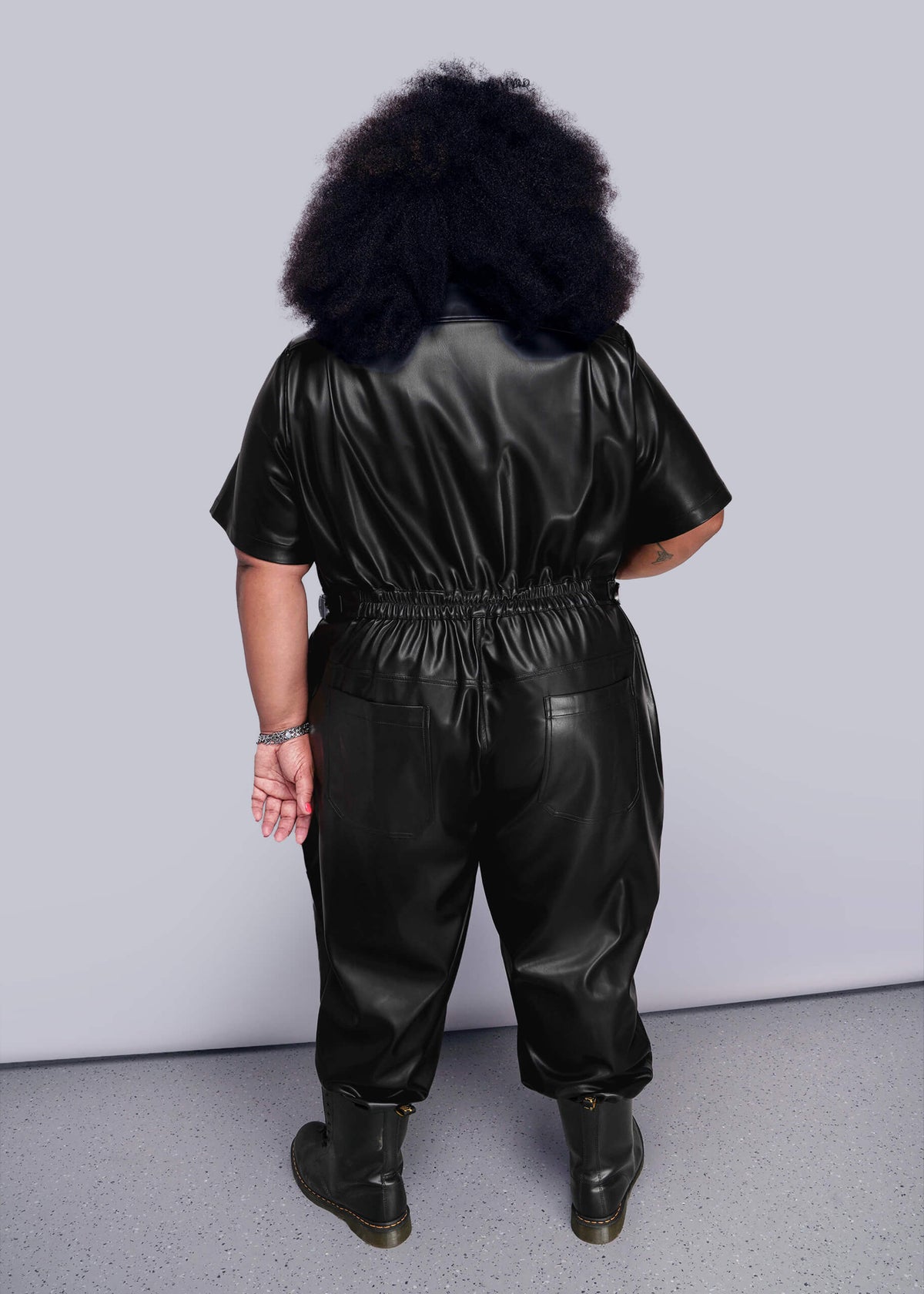 1X=Person wearing a black vegan leather jumpsuit against a plain background