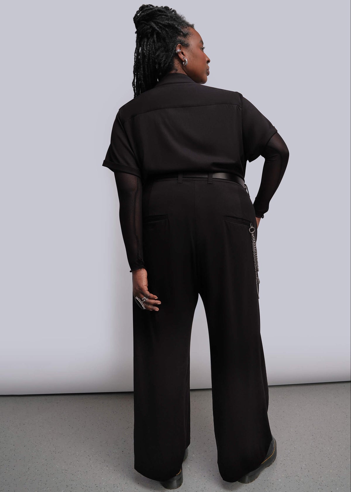 1X=A model shown from the back wearing a black wide-leg jumpsuit with a black belt and a silver wallet chain detail, set against a plain light gray studio background.