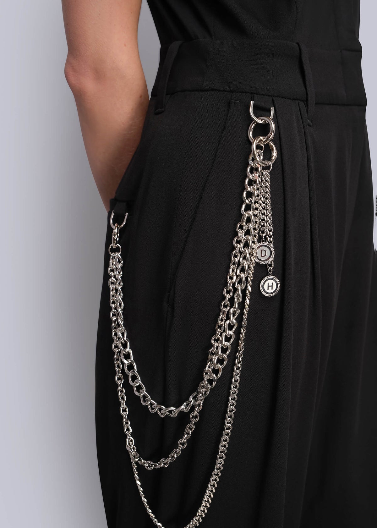 S=A close-up of a person wearing black high-waisted jumpsuit with a long silver wallet chain accessory hanging from the waistband, featuring interlocking circular rings and a small round logo charm.