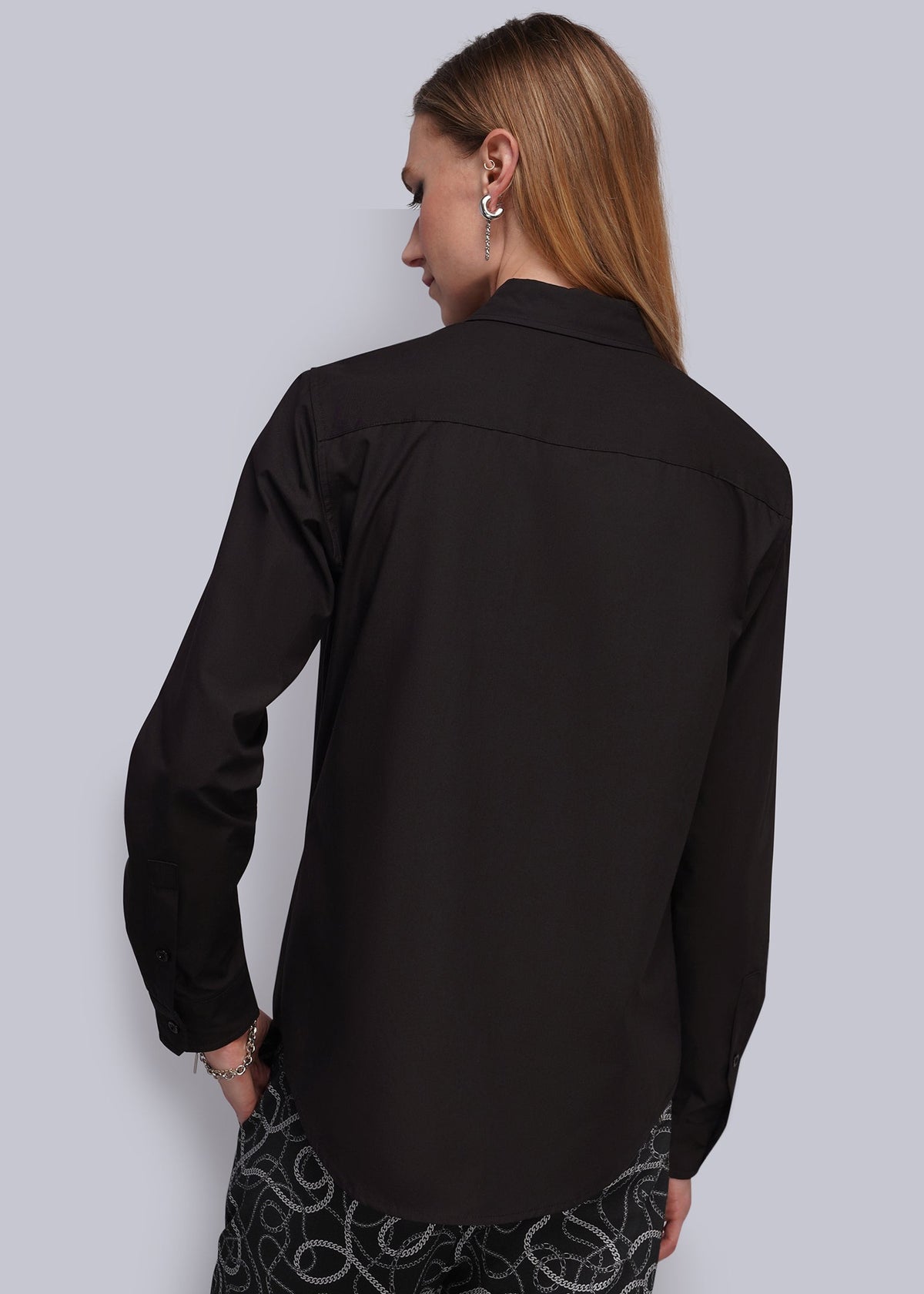 S=A woman shown from the back wearing a black long-sleeve button-up shirt and black pants with a white abstract pattern, set against a solid light gray background.