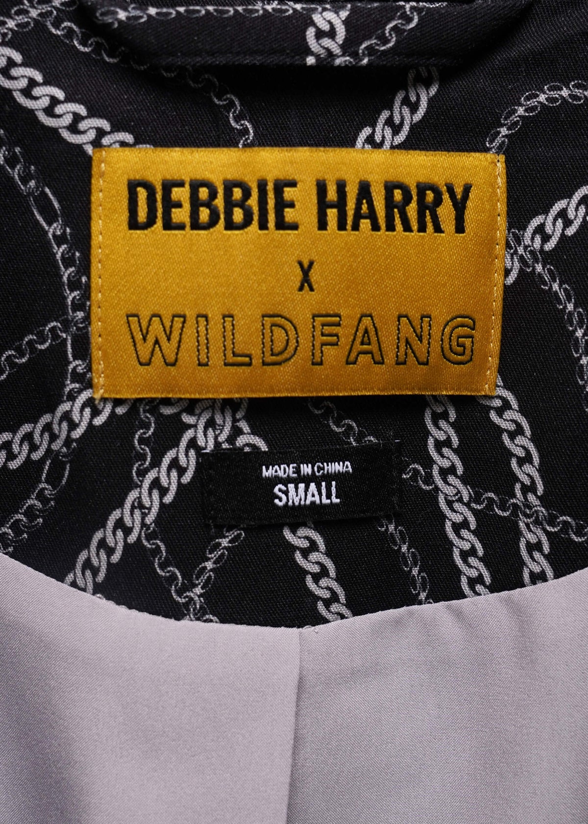 Close-up of a clothing label reading 'DEBBIE HARRY x WILDFANG' in black text on a mustard yellow patch, with a black fabric background patterned with white chain lines.