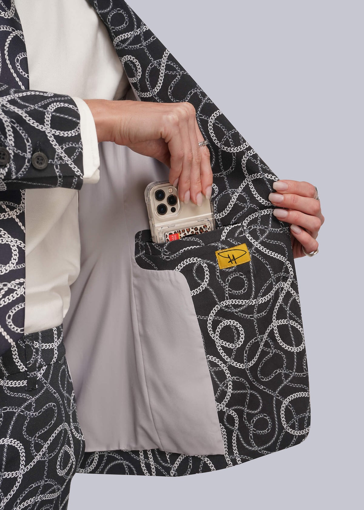 S=A close-up of a person wearing a black blazer with a white chain-link pattern, holding the lapel open to show a light gray interior pocket containing a smartphone and a small yellow tag.