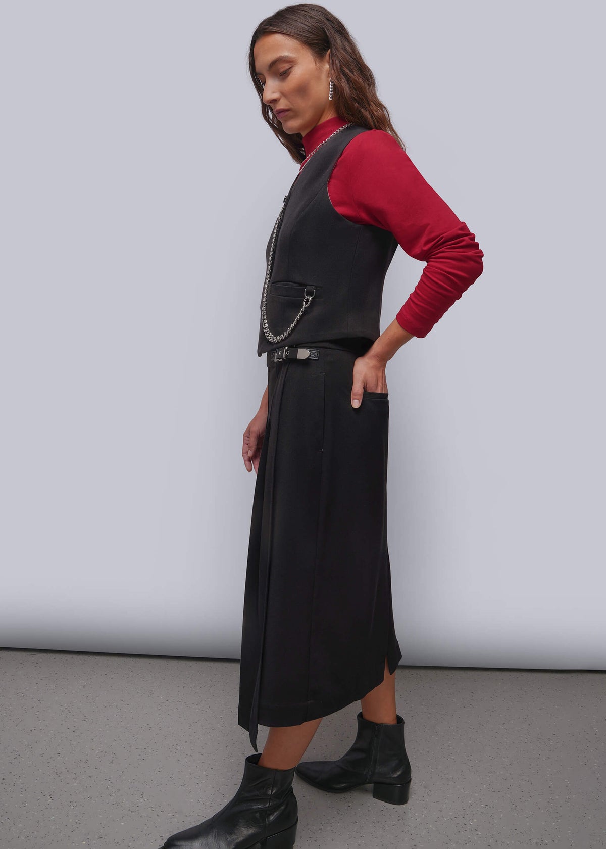S=A woman models a black vest with matchin midi skirt with a silver chain detail, layered over a red long-sleeve top and paired with black ankle boots, set against a plain studio background.