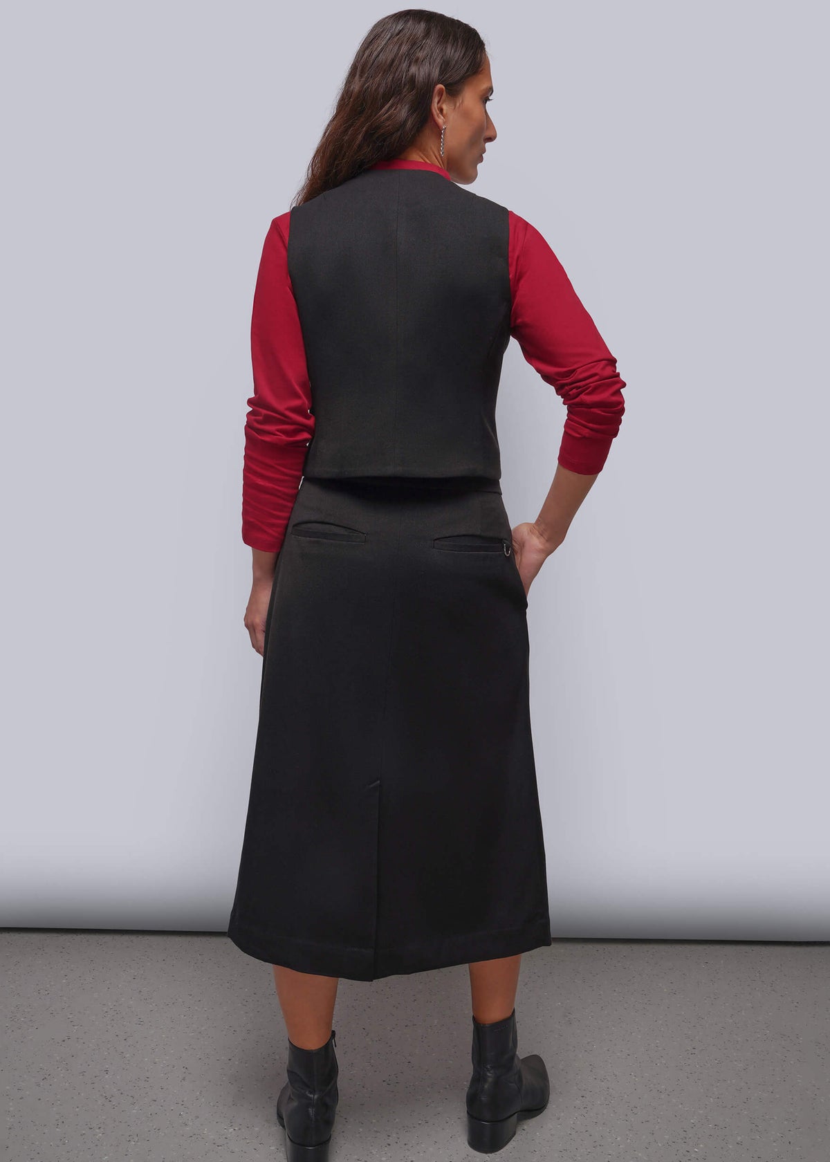 S=A woman is shown from the back wearing a black sleeveless vest and matching black midi skirt over a red long-sleeve top, paired with black ankle boots, against a plain light gray studio background.