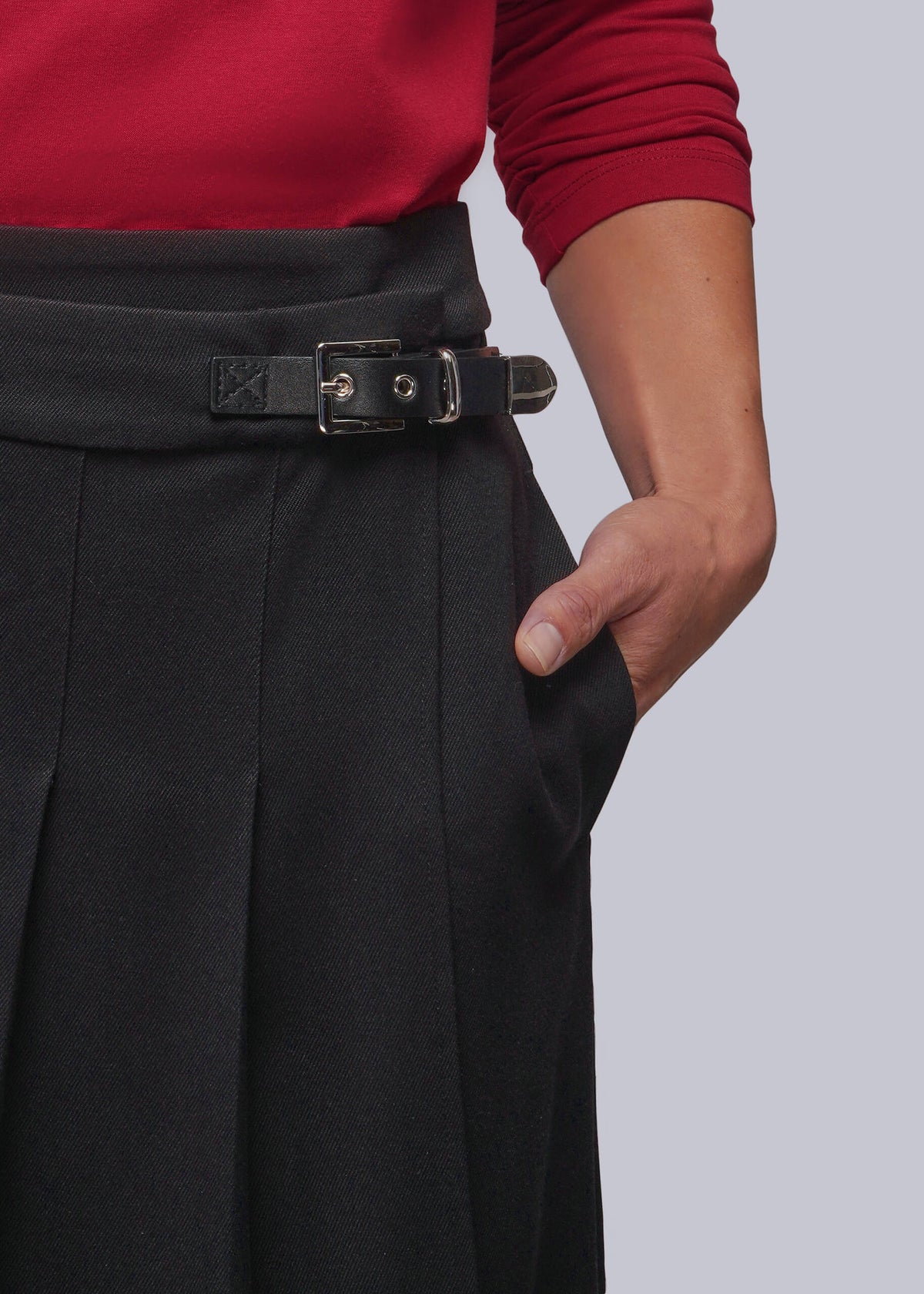 S=Close-up of a person wearing black pleated skirt with a black belt strap closure with silver buckle, paired with a red long-sleeve top, against a light gray background.