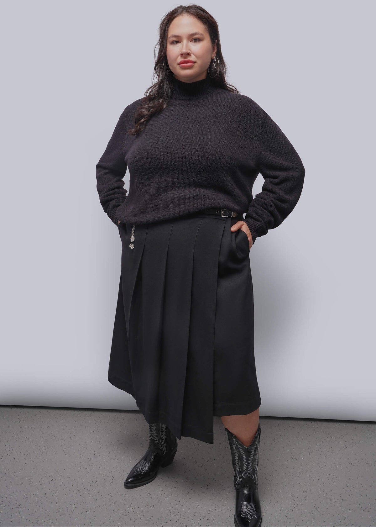 1X=A woman modeling a black turtleneck sweater and a matching dark pleated midi skirt, paired with black western boots, against a light gray studio background.