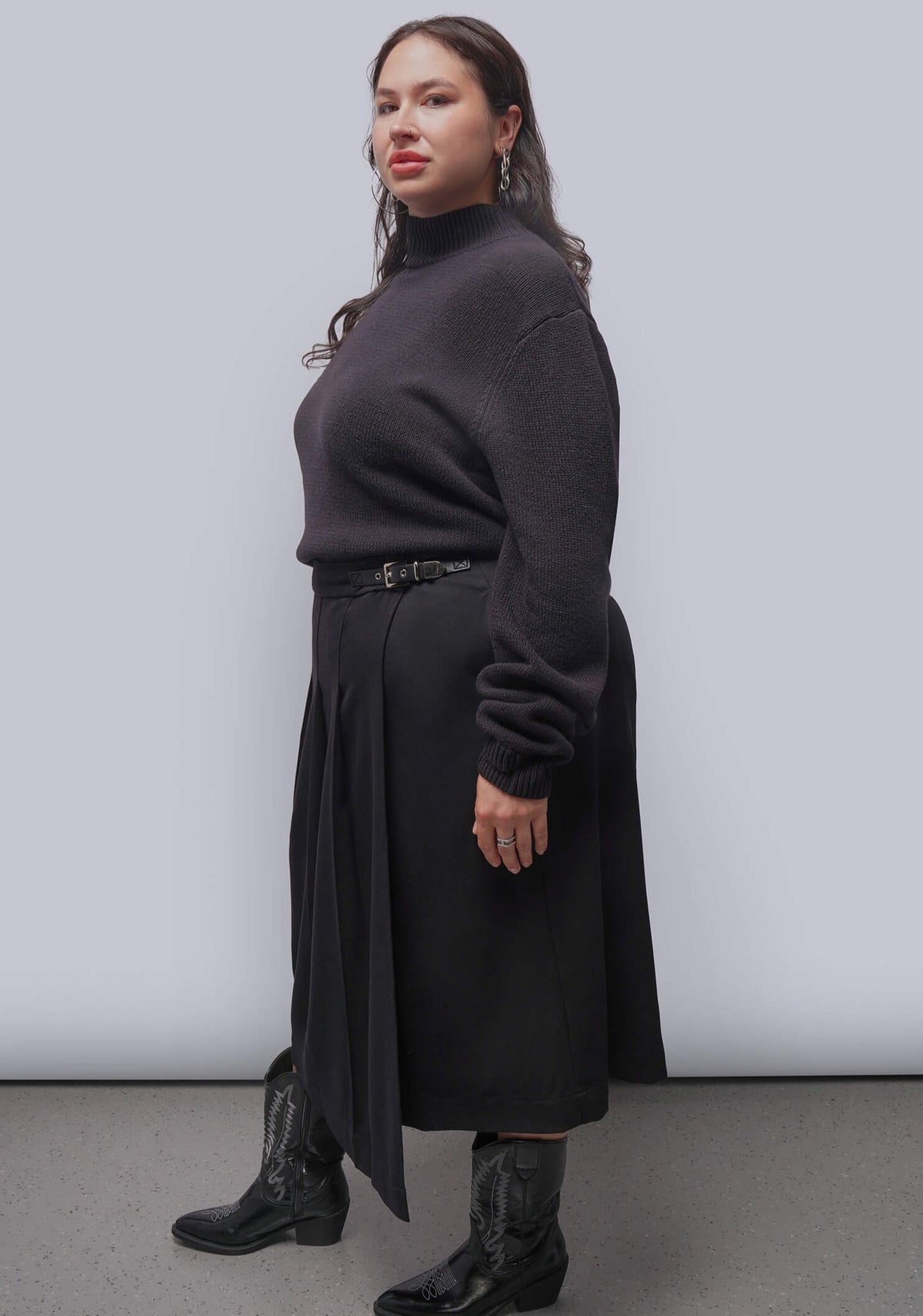 1X=A woman modeling a black knit mock-neck sweater paired with a black skirt and black leather boots, standing against a light gray studio background.