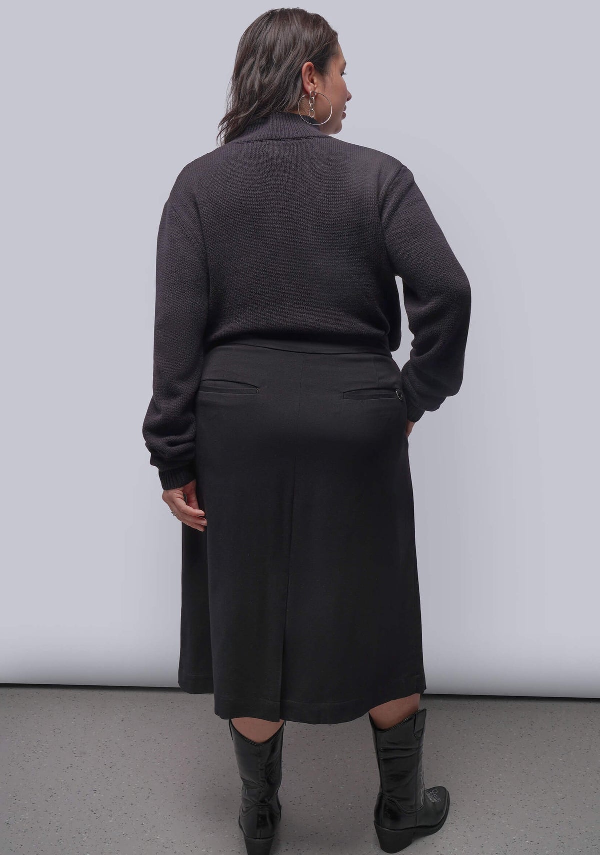 1X=A woman viewed from the back wearing a black knit long-sleeve top and a matching dark midi skirt, paired with dark gray knee-high boots, against a light gray studio background.