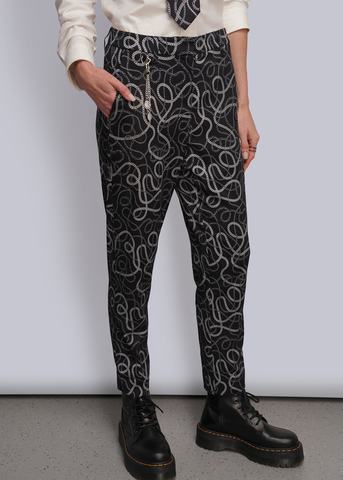 S=A person wearing black pants with a white and grey chain-link wavy pattern, styled with a cream shirt, a patterned tie, and silver chain accessories, paired with black leather boots.