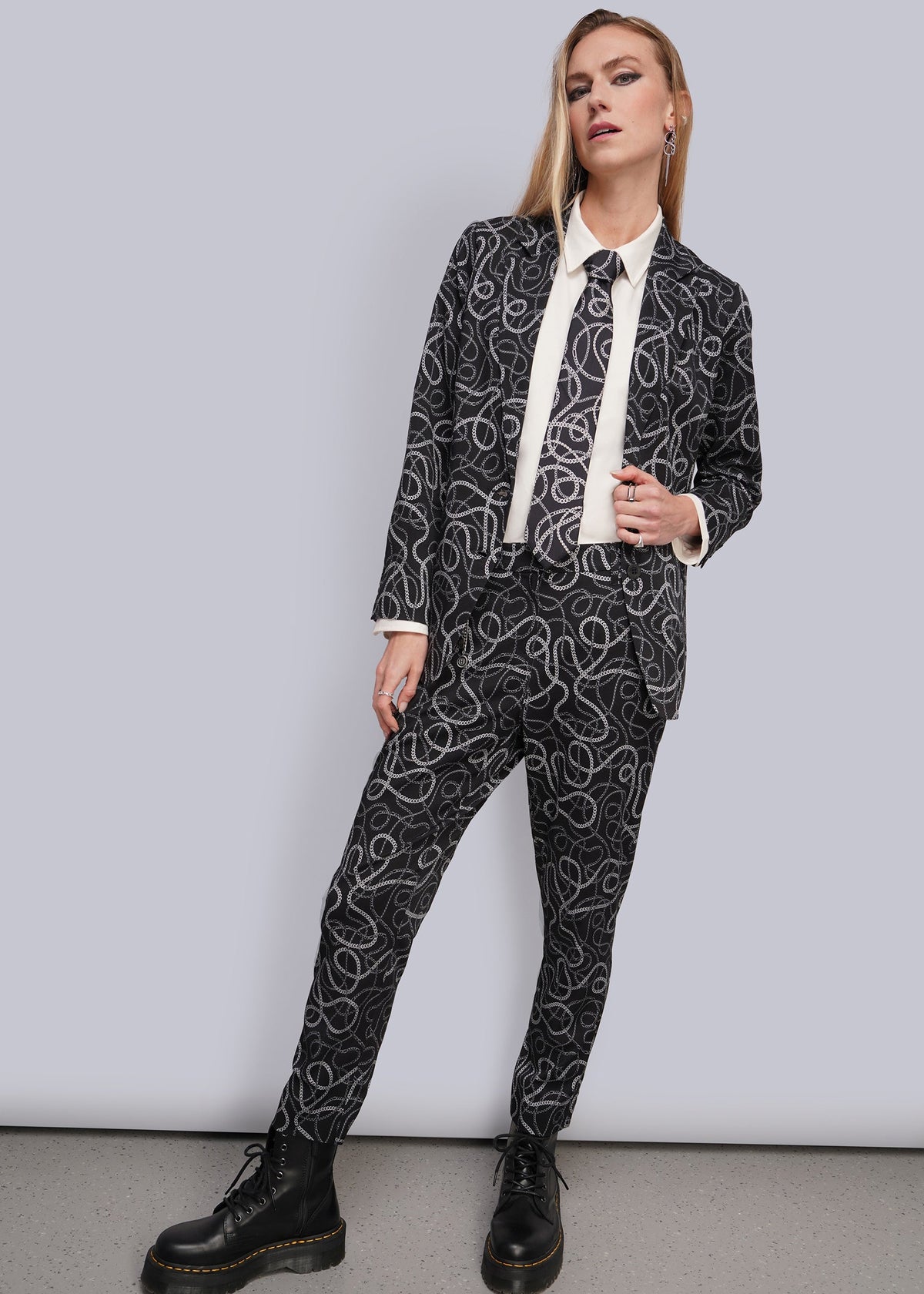 S=A woman modeling a black and white chain-link patterned suit with a white shirt and black boots, standing against a light gray studio background.