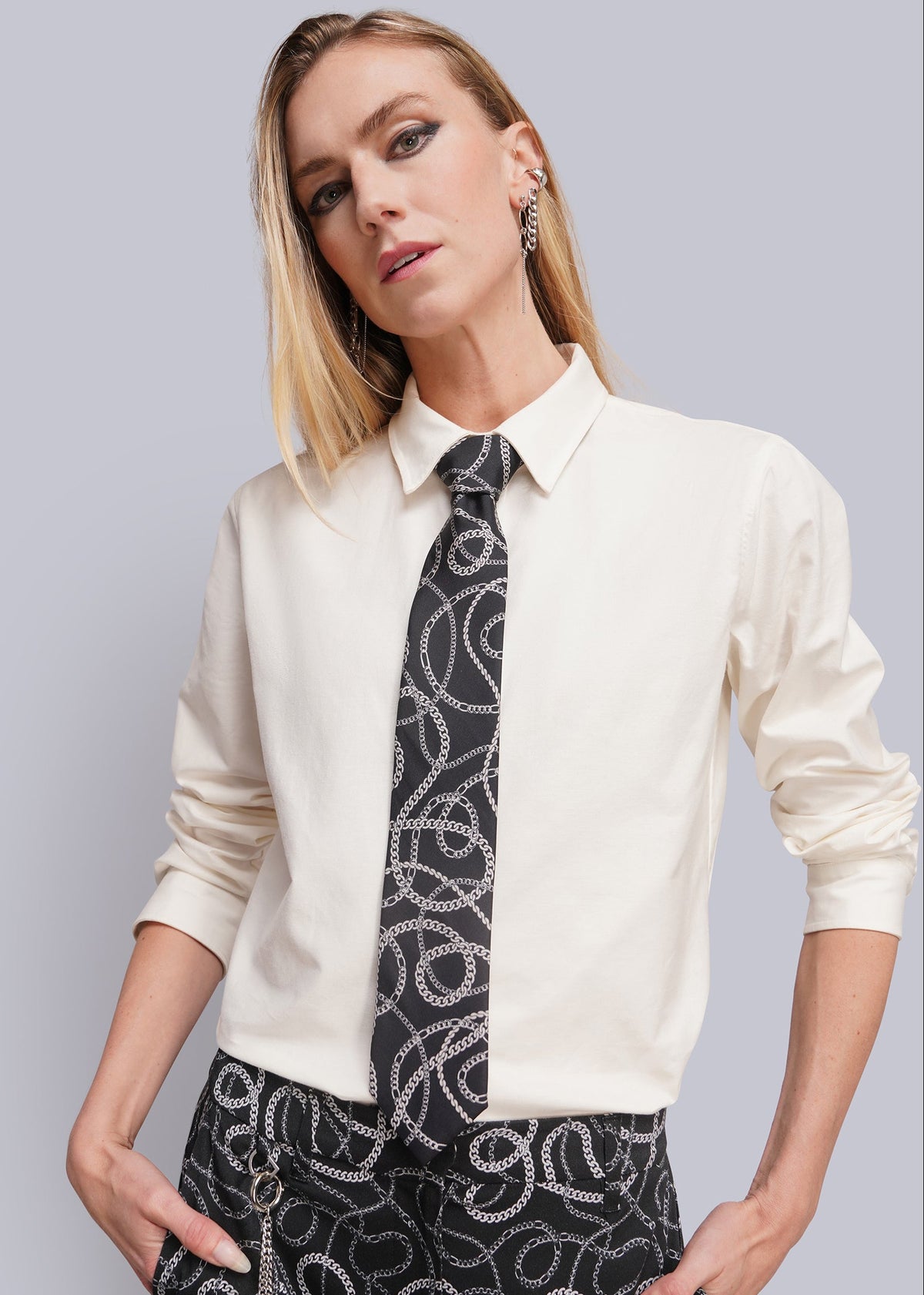 A woman modeling a cream-colored long-sleeve collared shirt paired with a black and white patterned tie and matching patterned trousers, set against a solid light gray background.