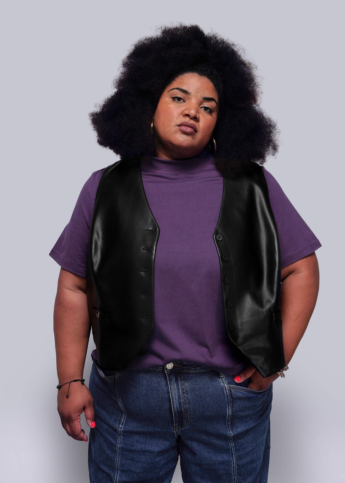 1X=Person wearing a purple shirt, black vegan leather vest, and blue jeans against a light gray background