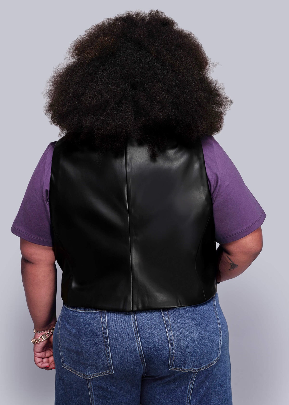 1X=Back view of person wearing a black vegan leather vest over a purple shirt with a plain background