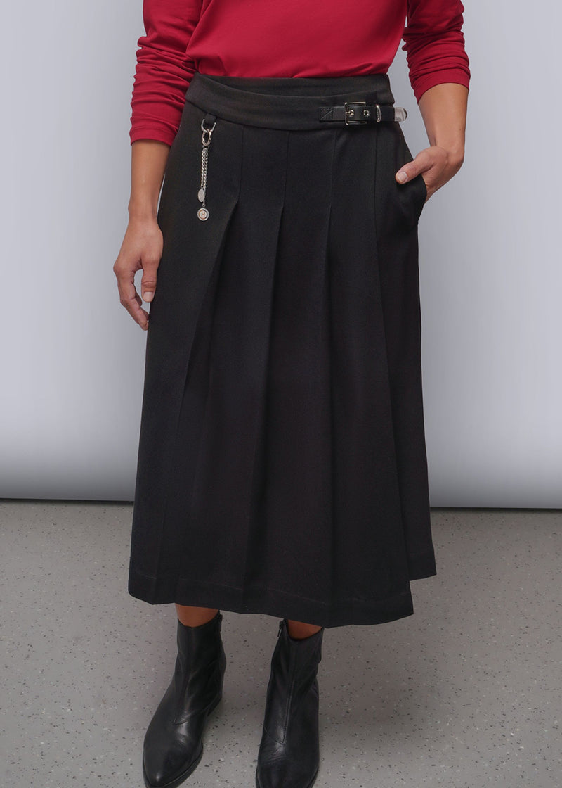 S=A person wearing a black pleated midi skirt with a matching belt and silver chain detail, paired with a red long-sleeve top and black ankle boots, standing against a light gray studio background.