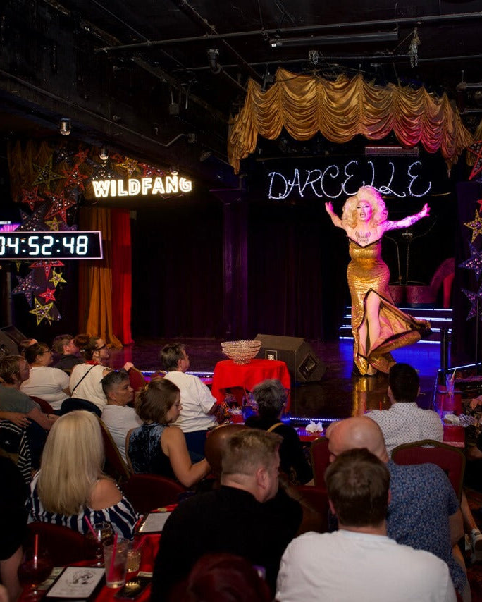 Drag queen in blonde wig and gold dress performs on stage at Darcelle's with crowd of seated people watching.