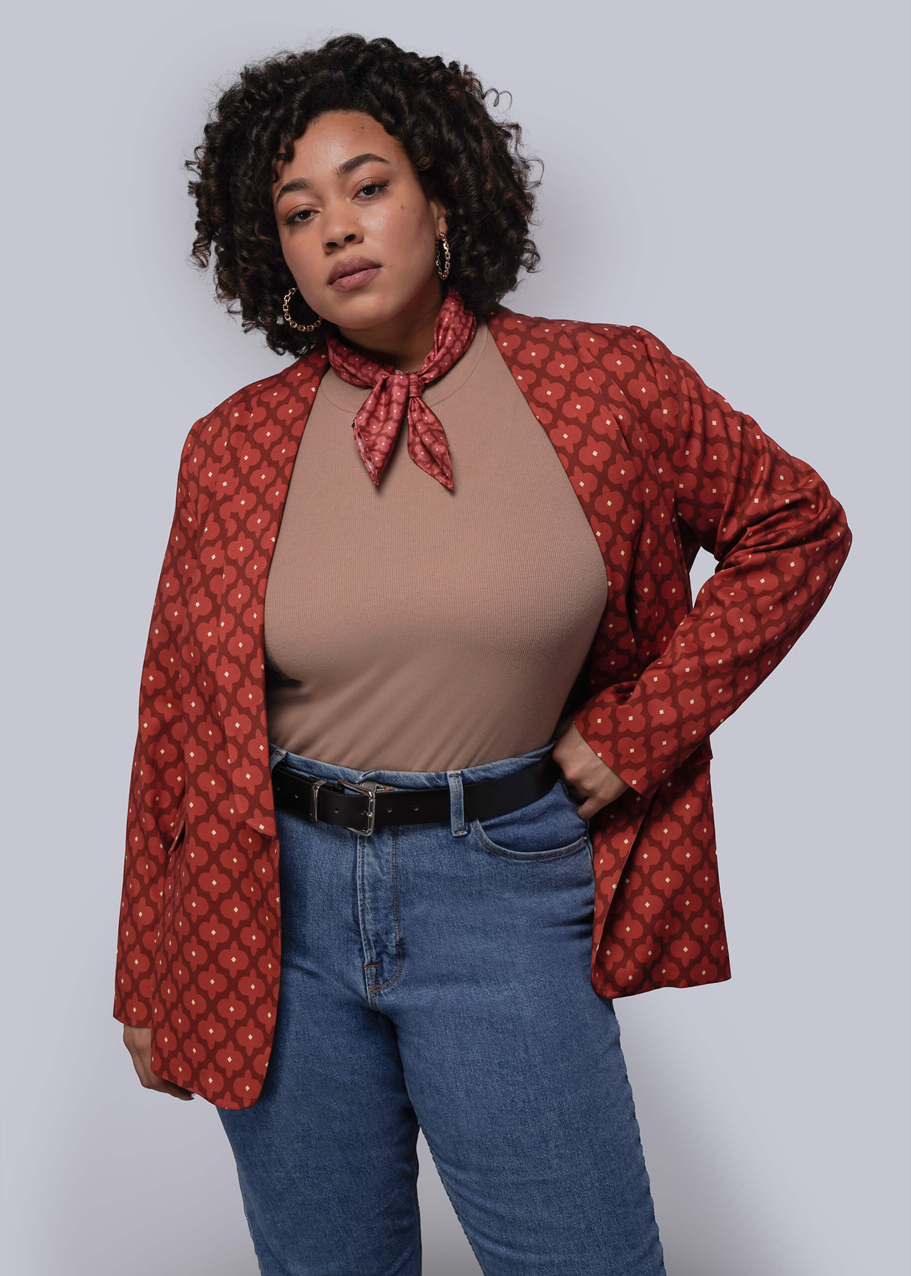 Person wearing a red patterned jacket over a brown top and blue jeans on a plain background