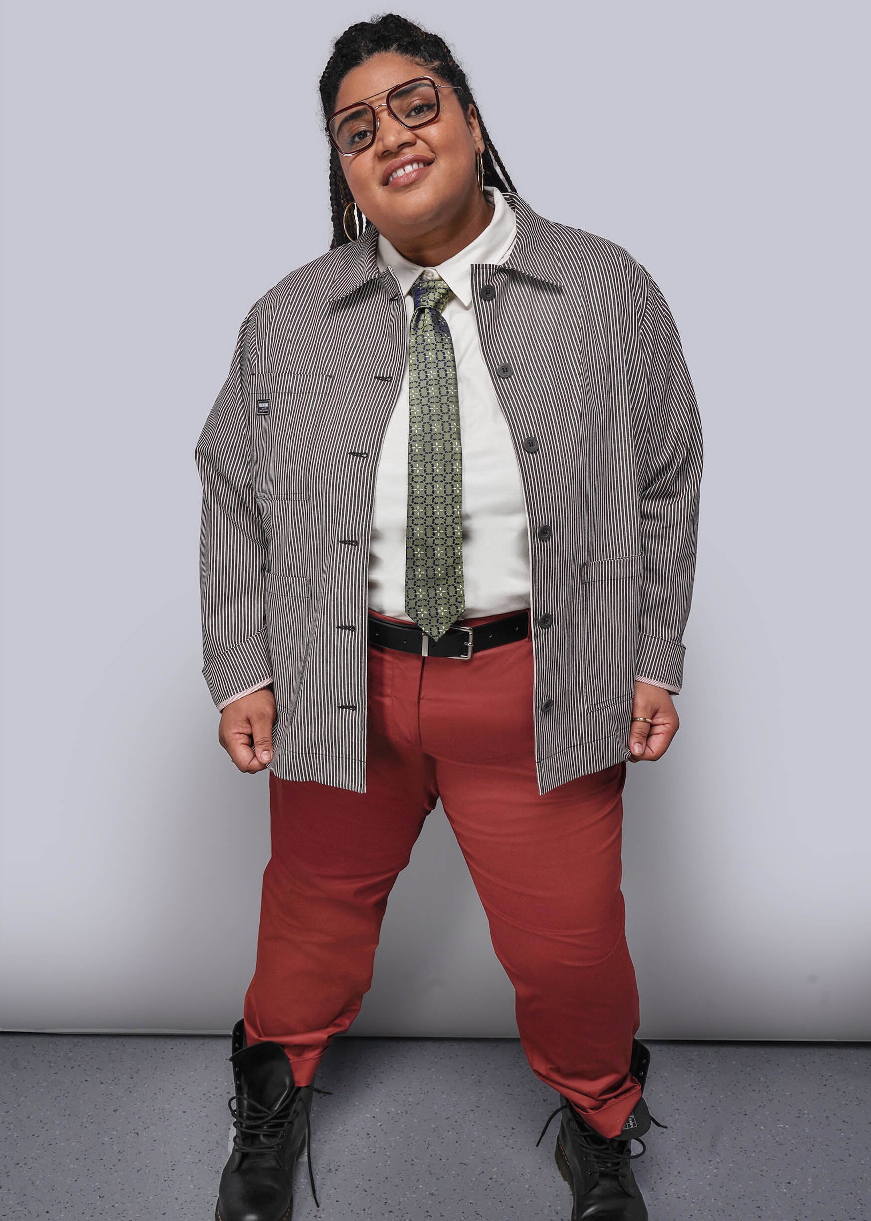 Person wearing a gray jacket, white shirt, red pants, and black shoes against a plain background
