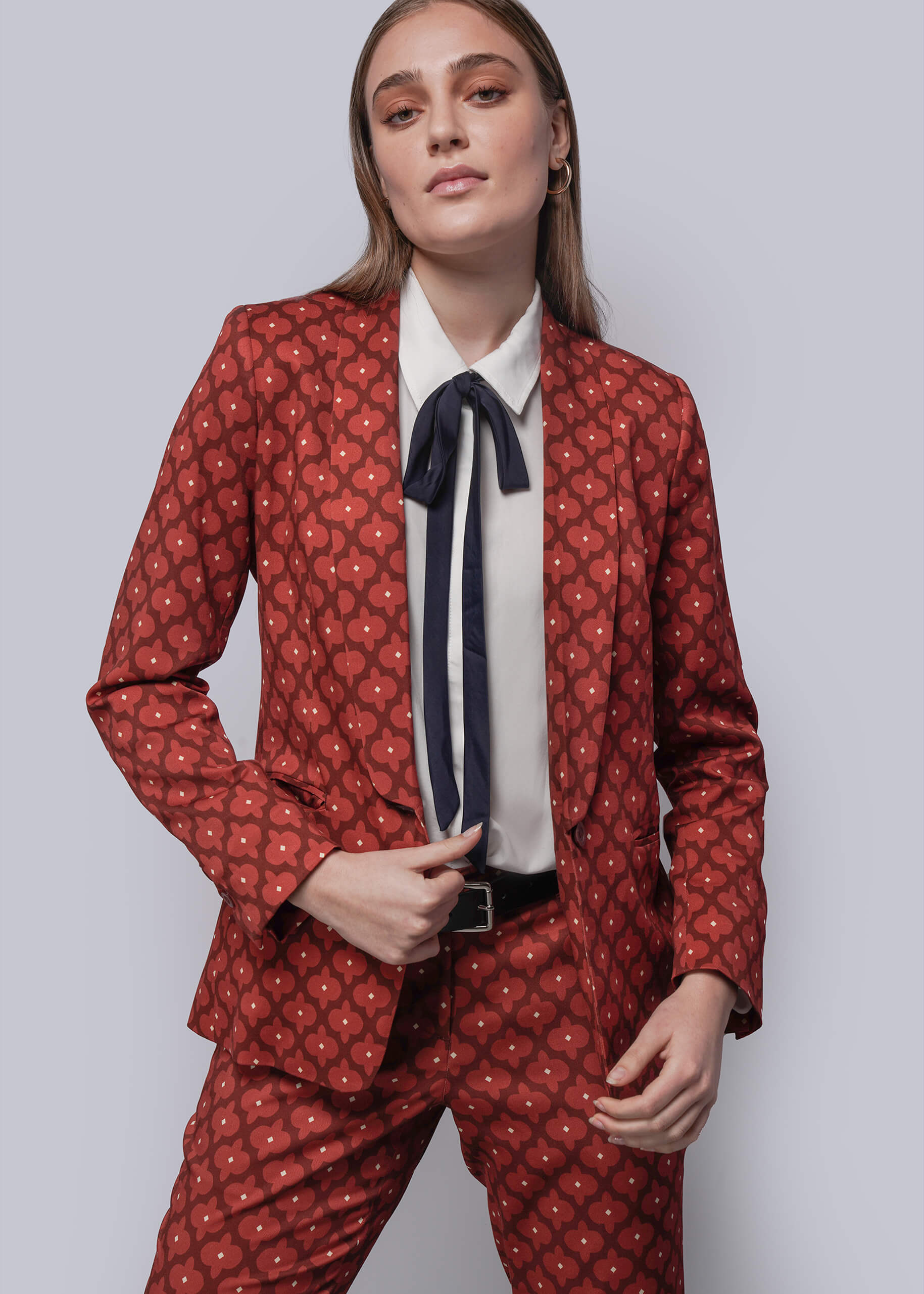 Woman wearing a red patterned suit with a white shirt with navy bow on a light gray background