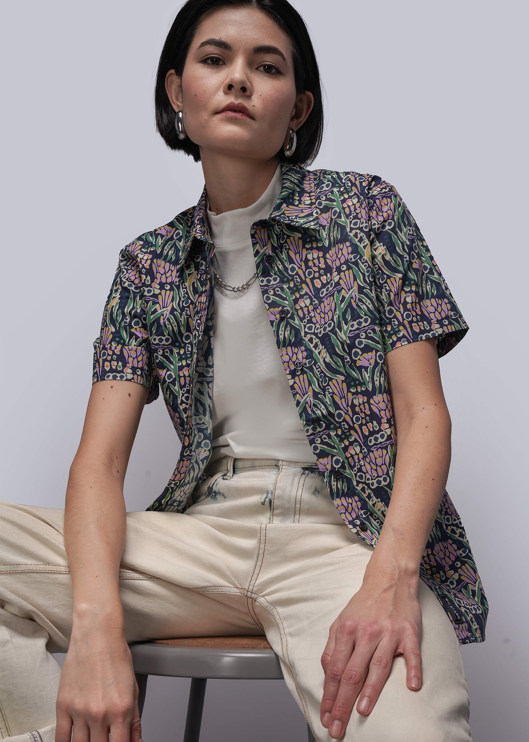Woman sitting on a chair wearing a floral shirt over a white top and beige pants.