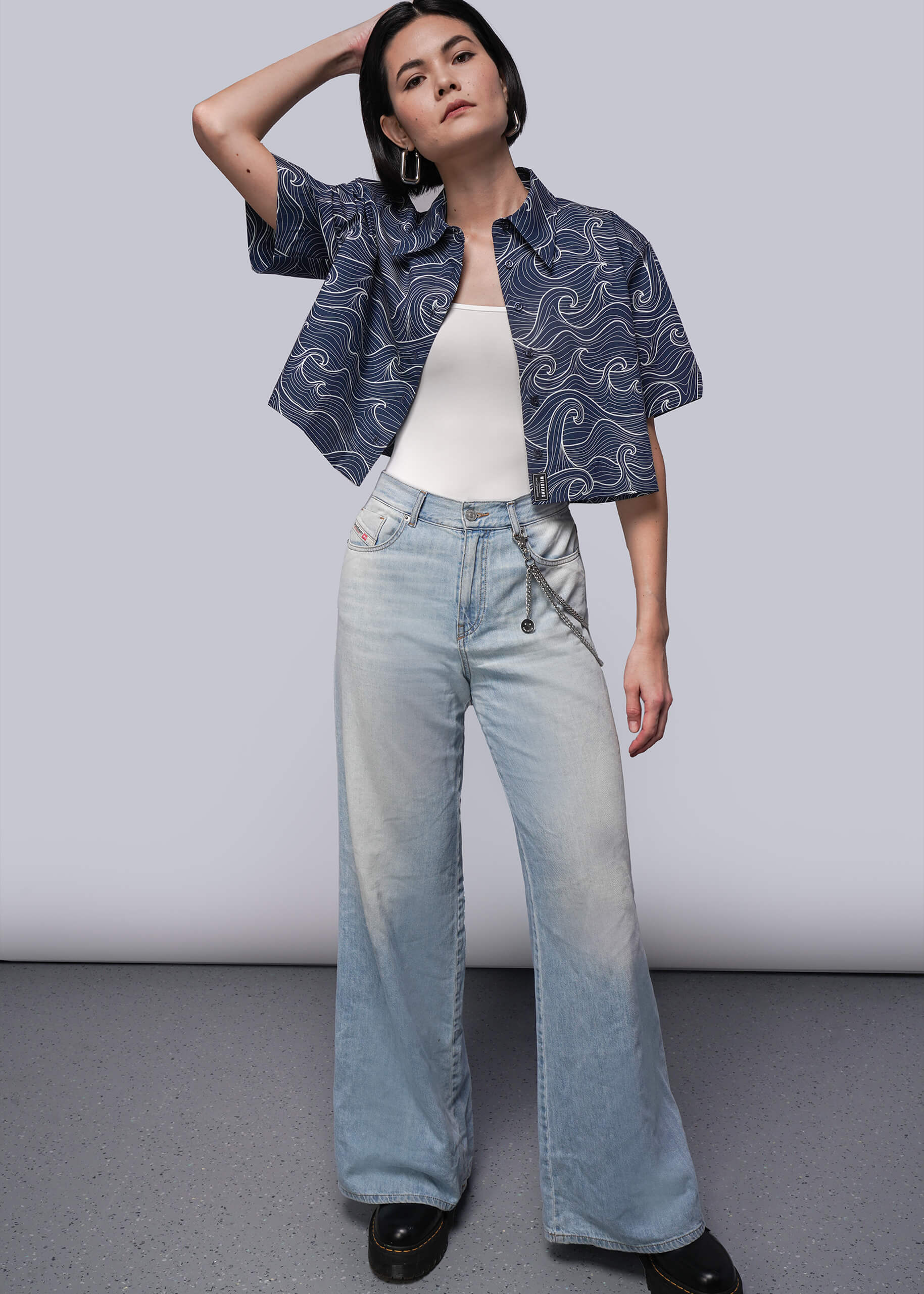 S=Woman wearing a patterned navy button up shirt, white top, and light blue jeans against a plain background