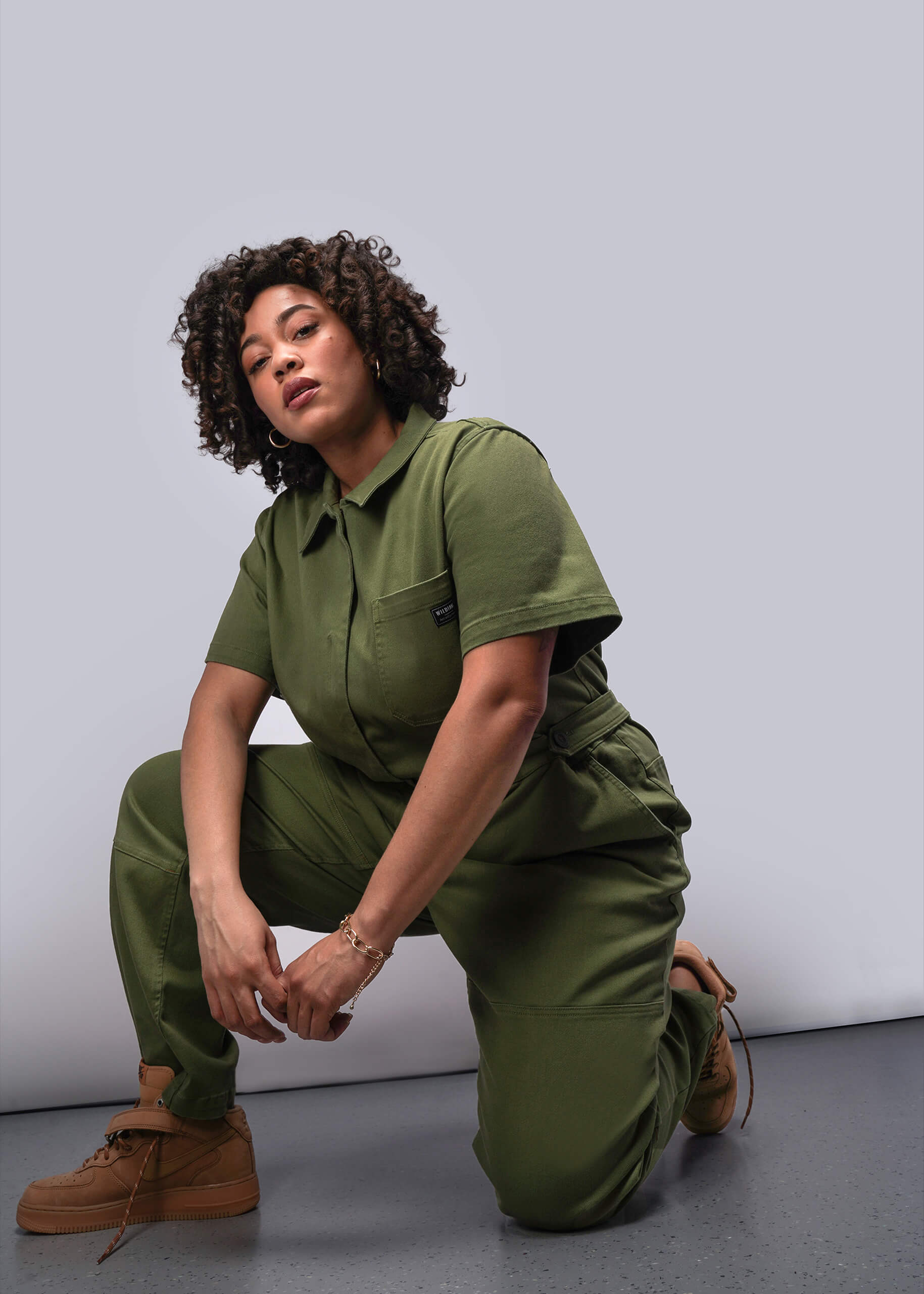 1X=Person wearing a green jumpsuit kneeling on the floor against a gray background