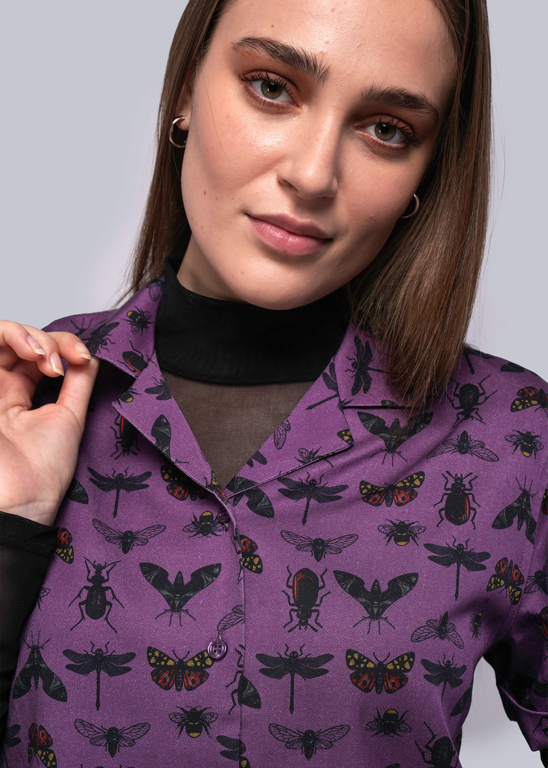 S=Woman wearing a purple shirt with insect pattern against a plain background