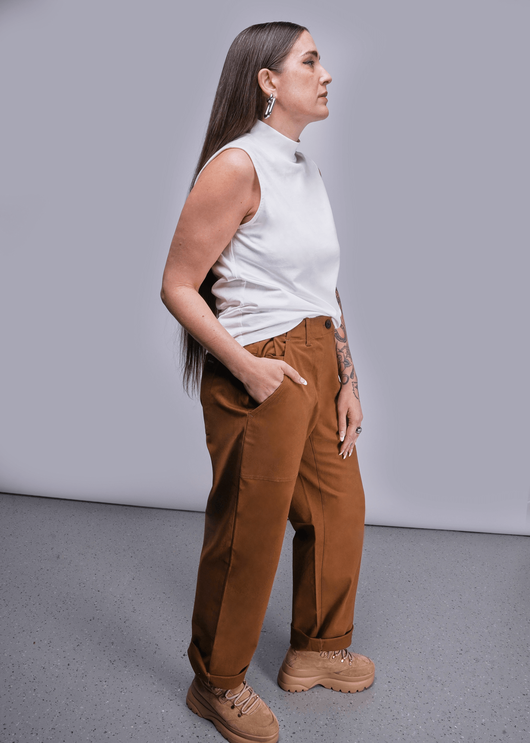 S=Side view of model with long brown hair standing in front of light grey background wearing The Everyday Mock Neck Tank in White and The Essential Utility Pant in Walnut with hand in pocket.