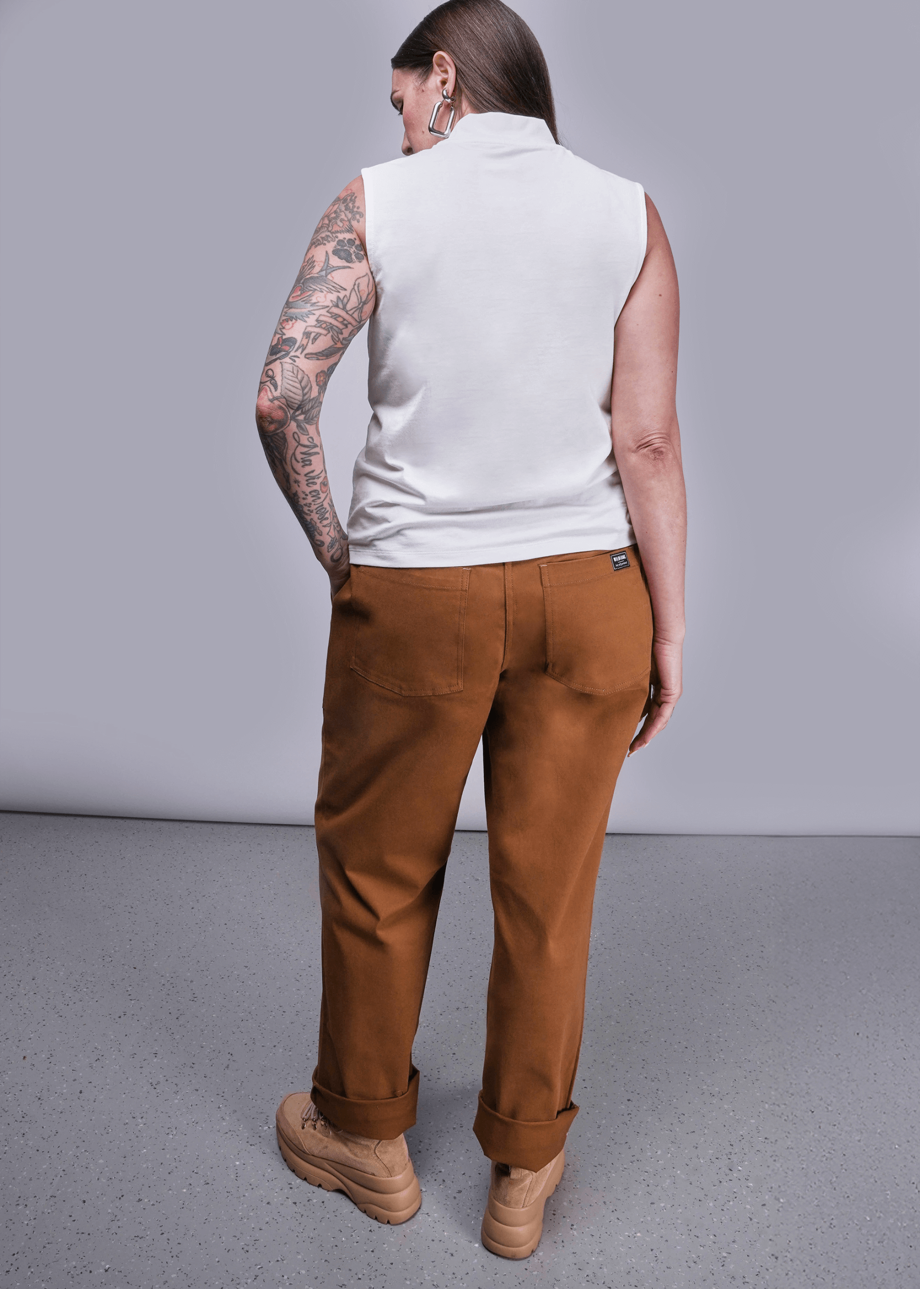 S=Back view of model with long brown hair standing in front of light grey background wearing The Everyday Mock Neck Tank in White and The Essential Utility Pant in Walnut with hand in pocket.