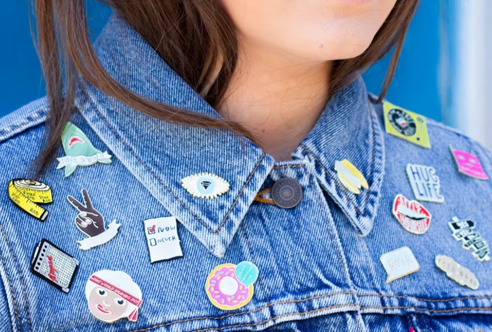 Close up of person wearing jean jacket with 16 different designed pins attached to it.