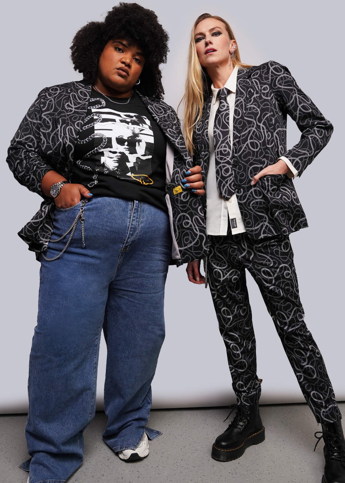 Two models posing in matching black and white abstract patterned suits, with one model wearing a black graphic t-shirt and blue jeans, and the other wearing a white collared shirt and tie.