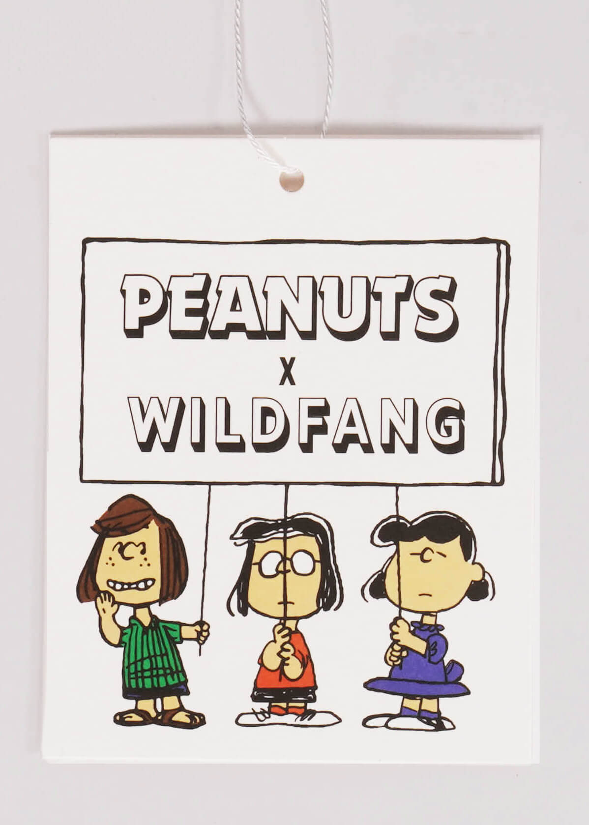 Hang tag laying flat on white background printed with Peanuts characters Peppermint Patty, Marcie, and Lucy holding sign that has the PEANUTS X WILDFANG logo on it.